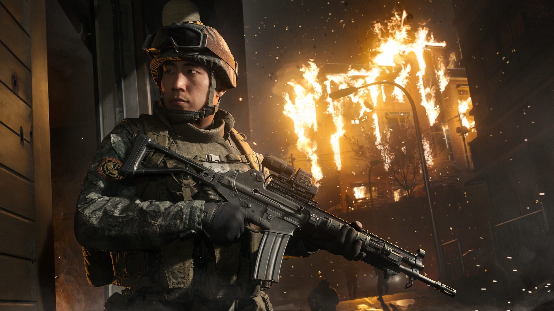 A soldier in a helmet, goggles and an assault rifle leans out from a corner while a building burns behind him on a night street.