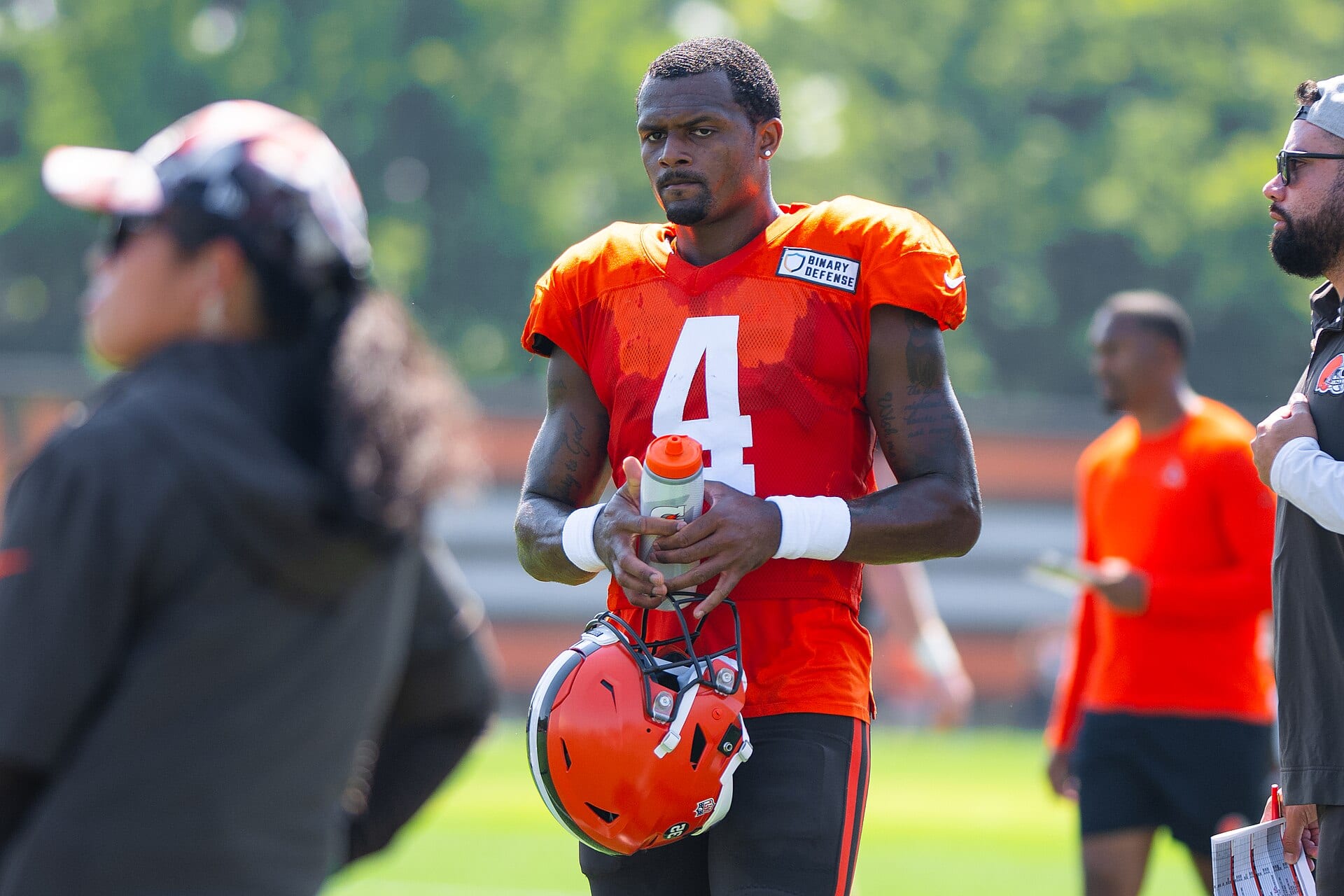 Un quarterback con camiseta naranja y el número 4 sostiene el casco y una botella de agua durante un entrenamiento.