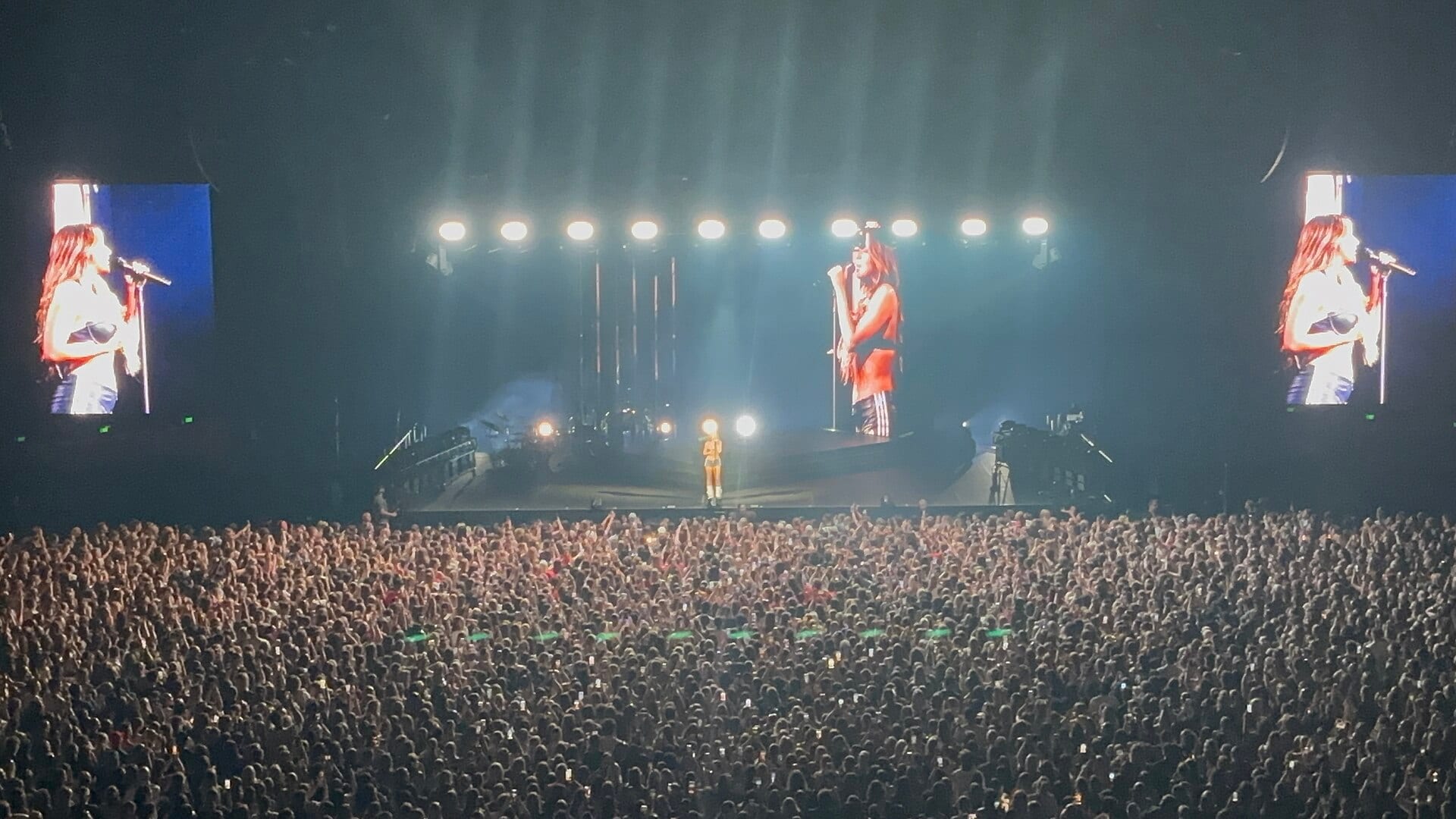 Singer Tate McRae performs alone on a lit stage, shown on side screens, with a large crowd in front.