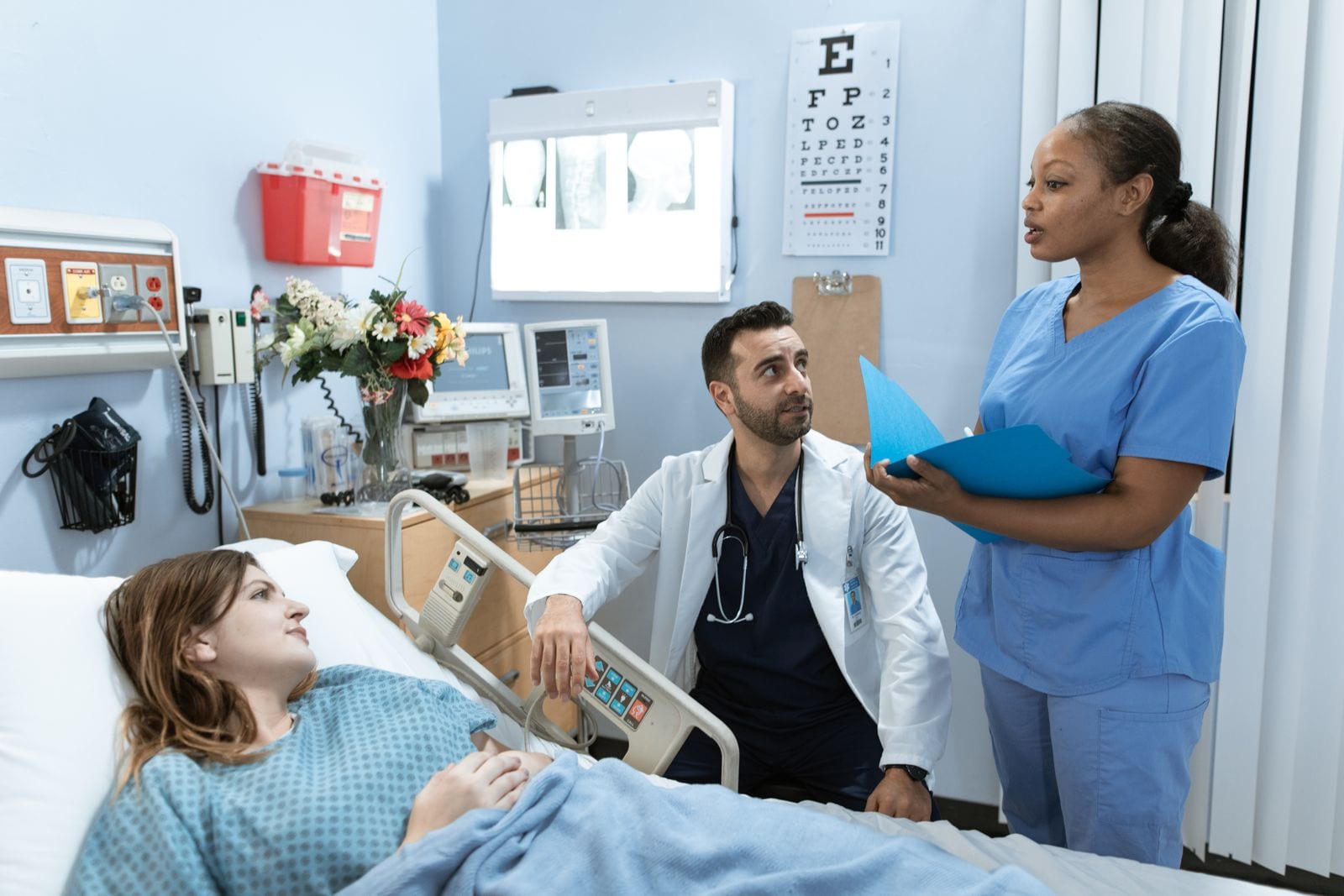 Un médico con bata blanca y una enfermera con una carpeta azul conversan junto a la cama de una paciente en una habitación de hospital.