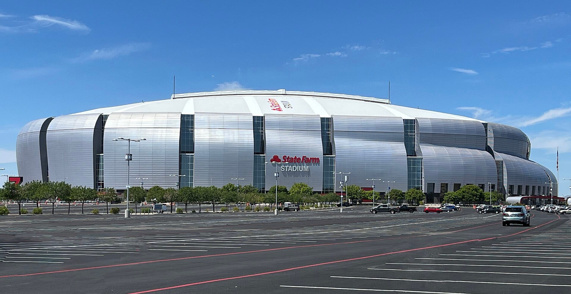 La fachada plateada del State Farm Stadium de Glendale vista desde un estacionamiento casi vacío, bajo un cielo azul.