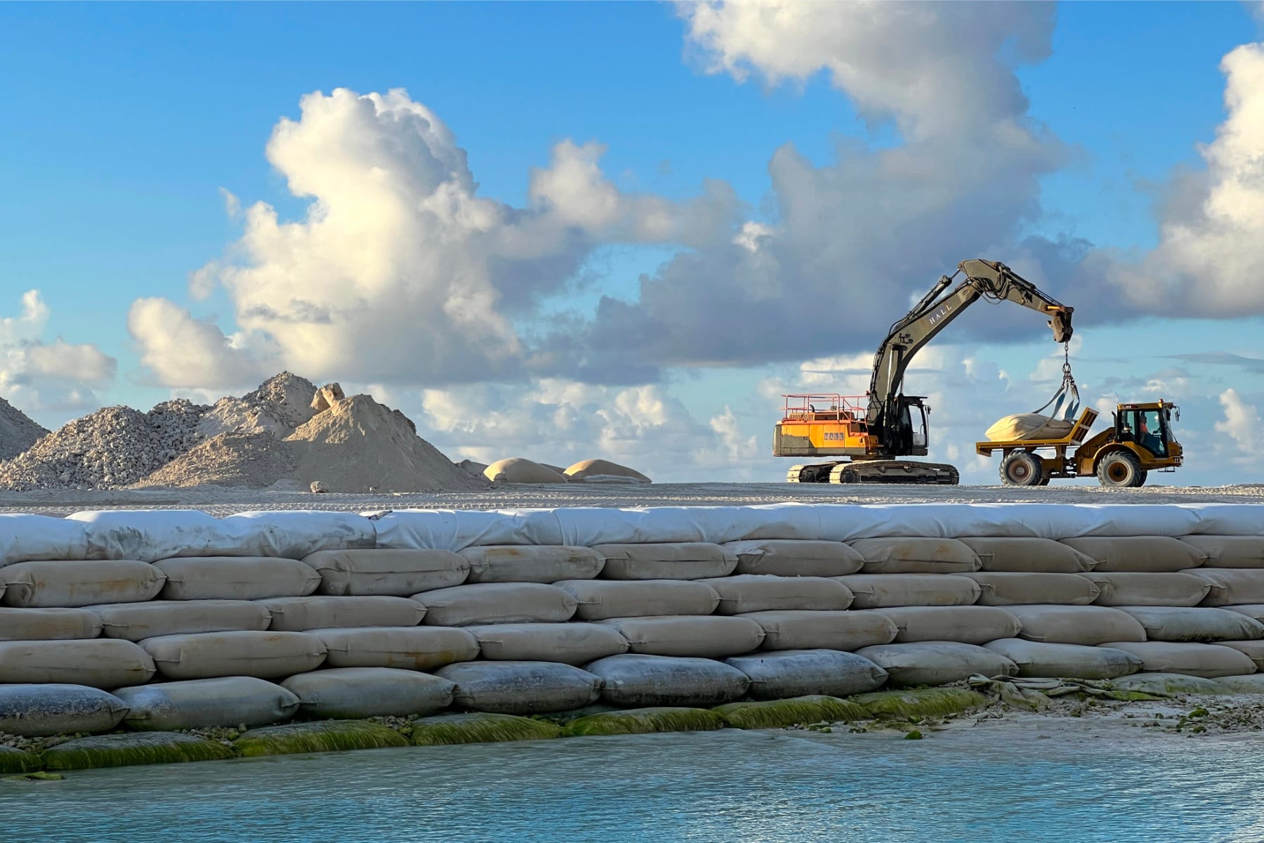 An excavator lifts a sandbag next to a wall of large geotextile bags, with the sea in the foreground.