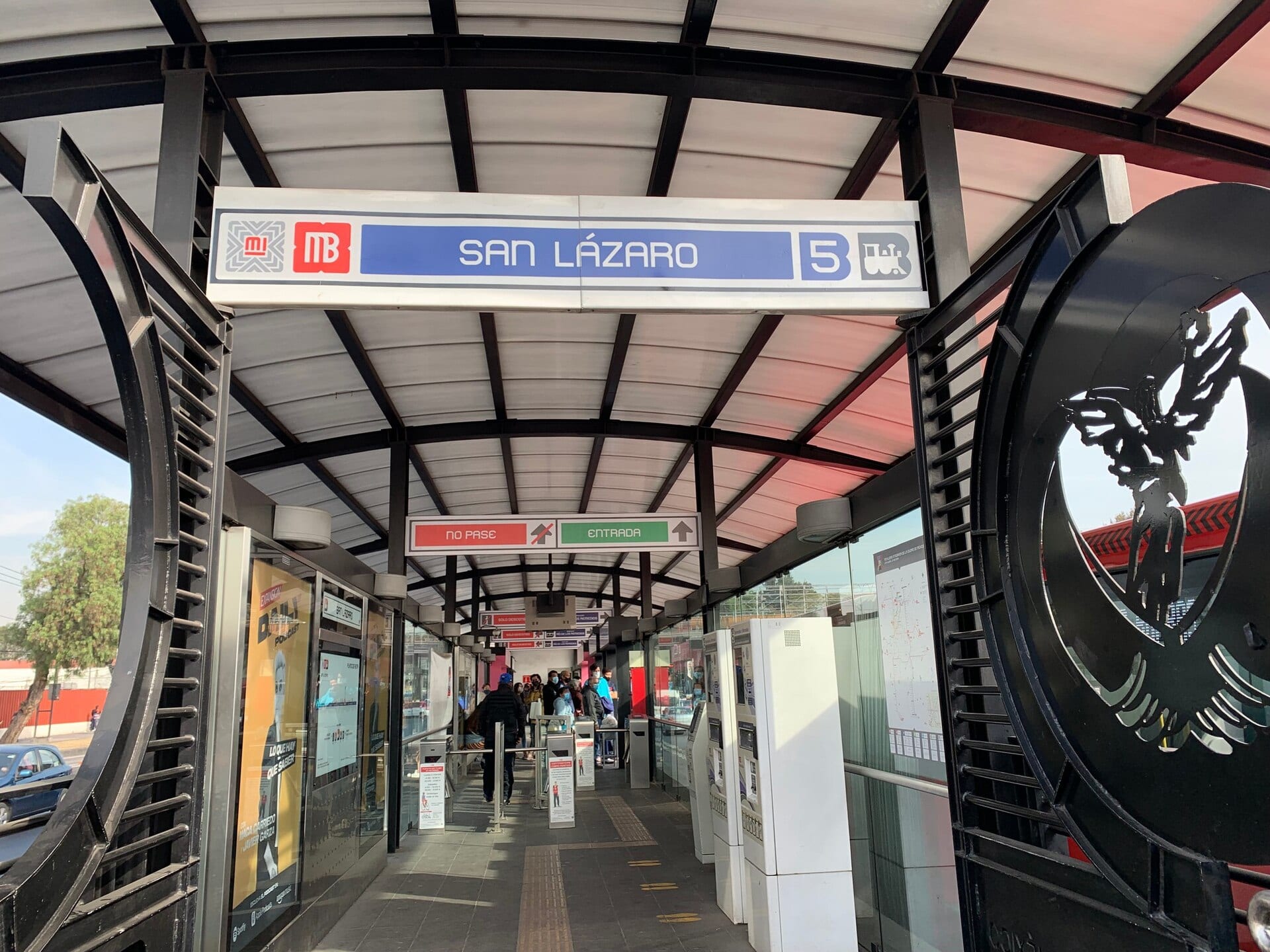 Entrada de una estación de la Línea 5 del Metrobús, con su letrero azul y blanco, torniquetes y pasajeros al fondo bajo una techumbre curva.
