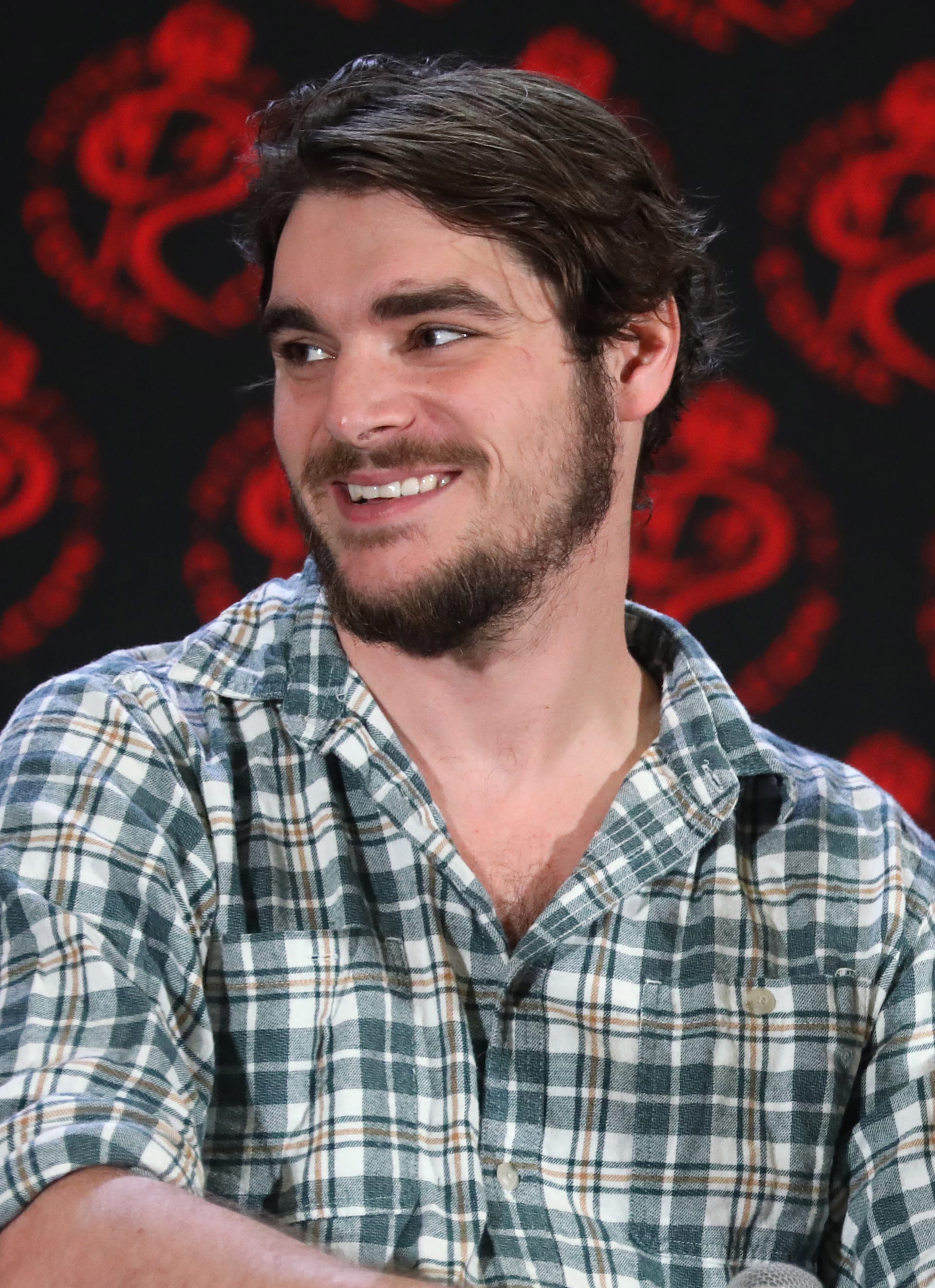 RJ Mitte, de cabello oscuro y barba, sonríe y mira hacia un lado con una camisa de cuadros frente a un fondo negro con motivos rojos.