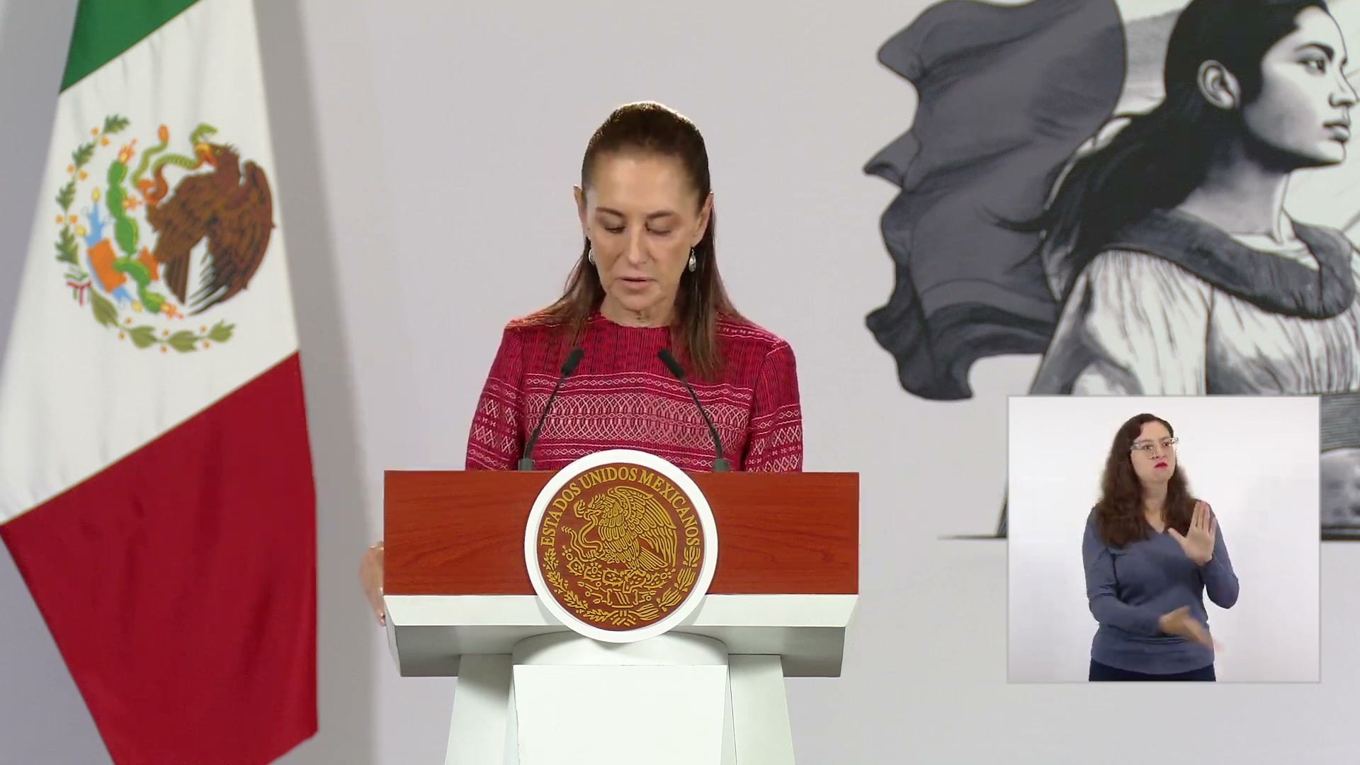 La presidenta Claudia Sheinbaum habla desde el atril con el escudo nacional, junto a una bandera y una intérprete de lengua de señas.