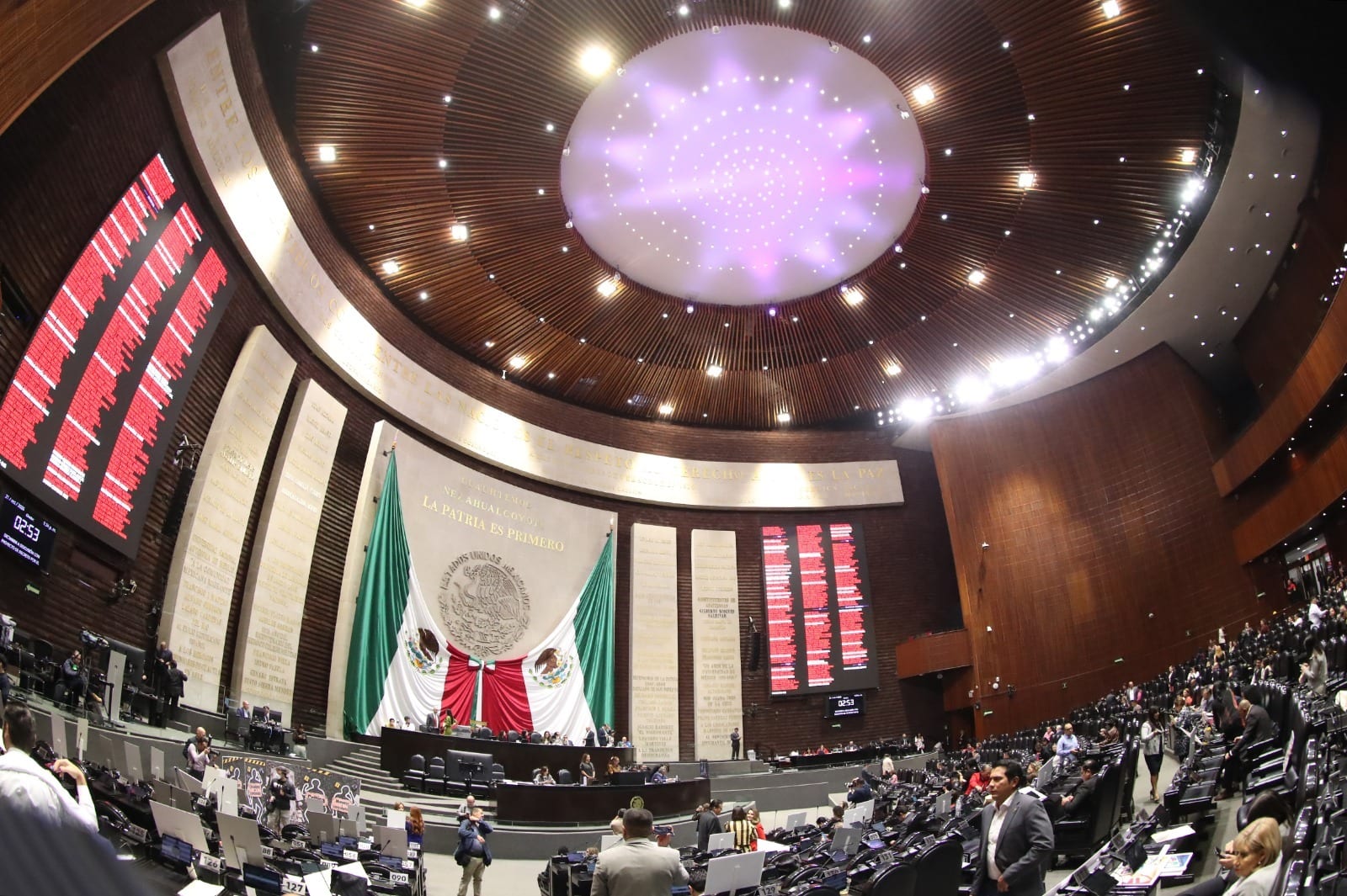 Salón de sesiones de San Lázaro visto con lente de ojo de pez, con los tableros de votación encendidos en rojo a ambos lados del muro con el escudo nacional y la bandera.