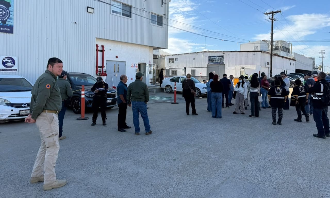 Personal de Protección Civil de uniforme verde y trabajadores esperan en un estacionamiento frente a una nave industrial blanca, con acordonamiento naranja.