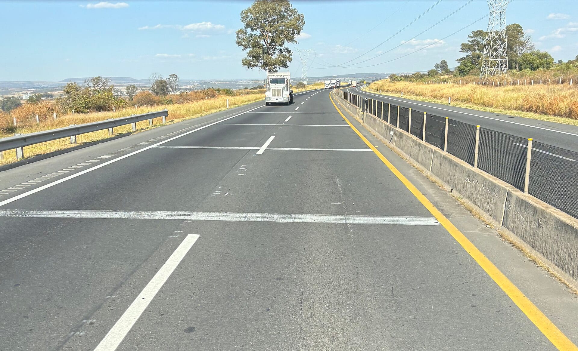 Autopista de varios carriles con tractocamiones blancos circulando al fondo, entre pastos secos y torres de electricidad.