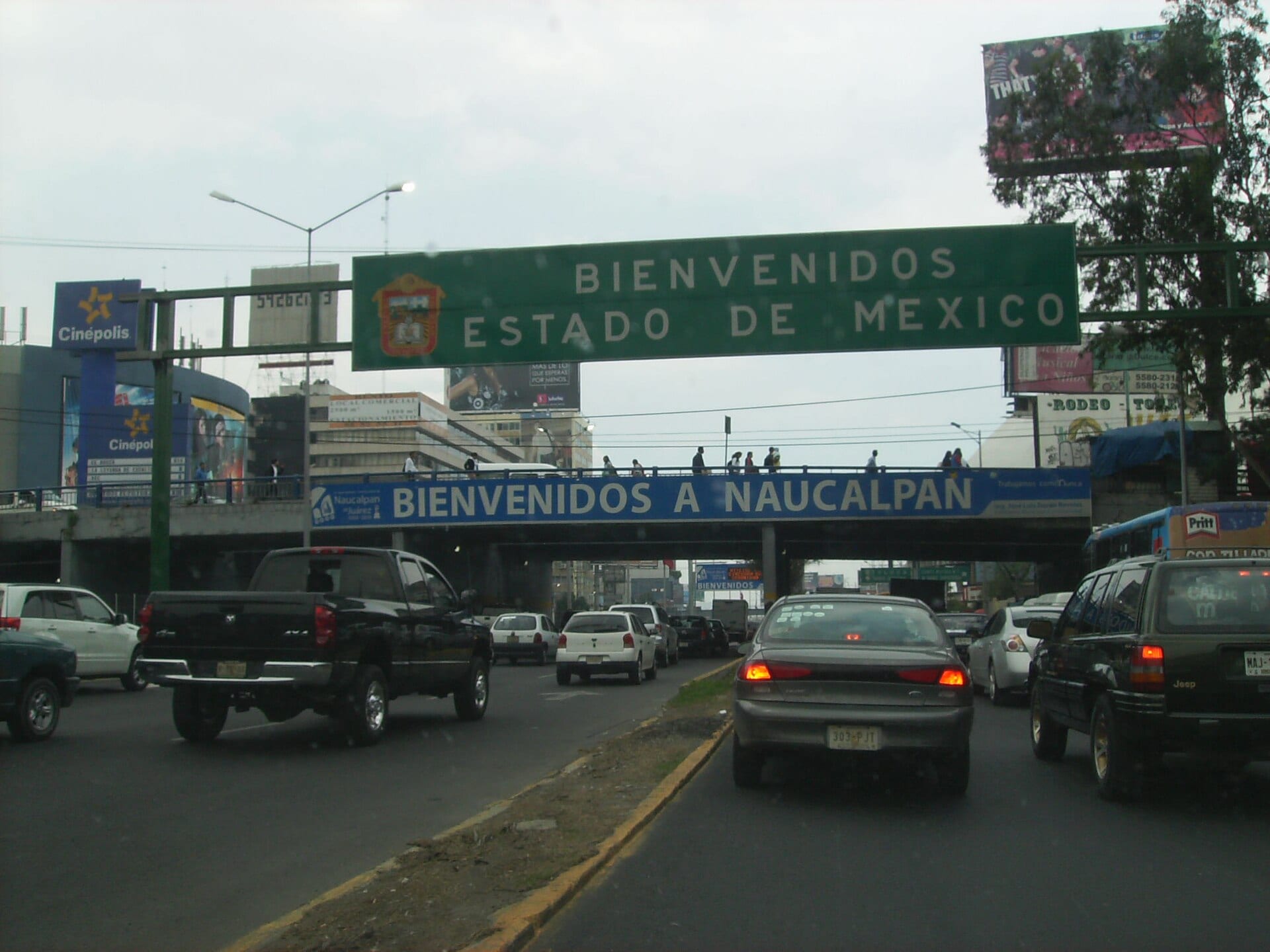 Vehículos avanzan en fila bajo un puente peatonal con un letrero que dice Bienvenidos a Naucalpan y otro verde de Bienvenidos Estado de México.