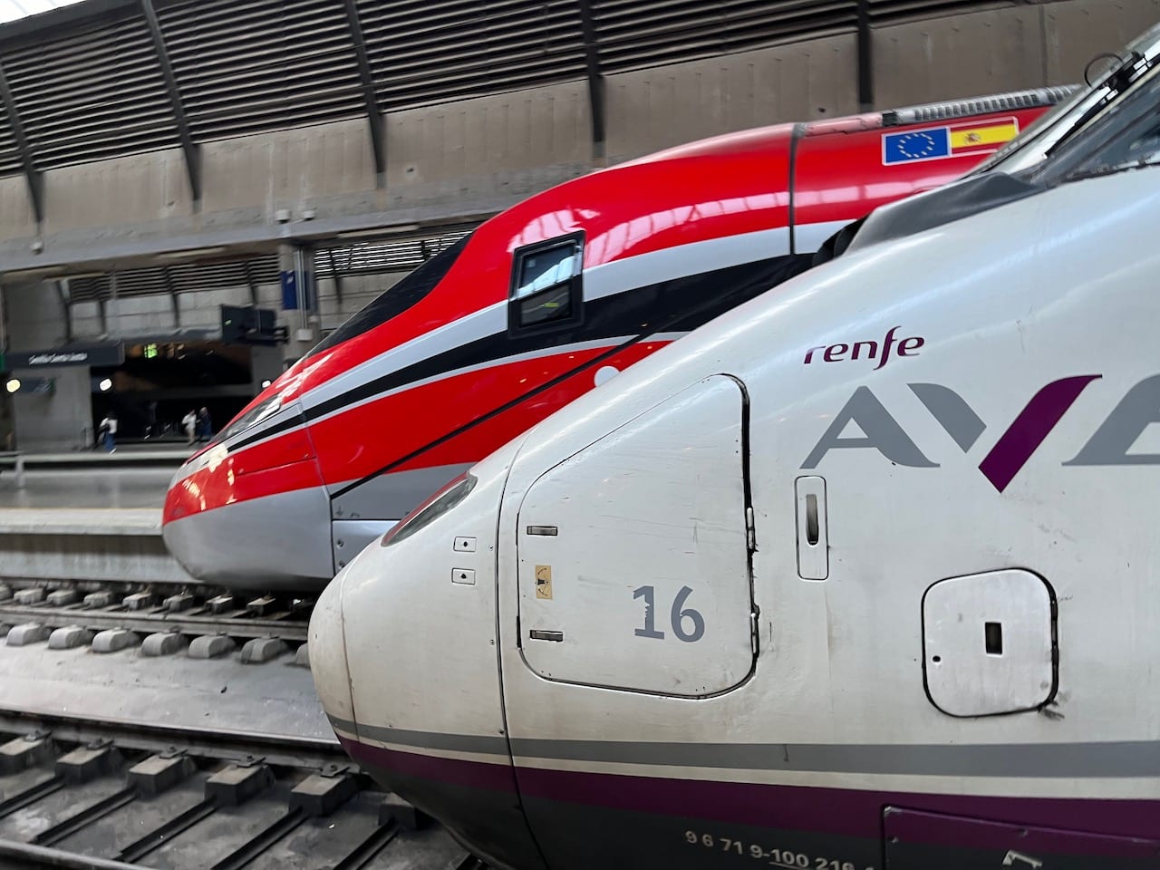 The noses of a white Renfe AVE train with purple lettering and a red Iryo train with black stripes, side by side at a station platform.