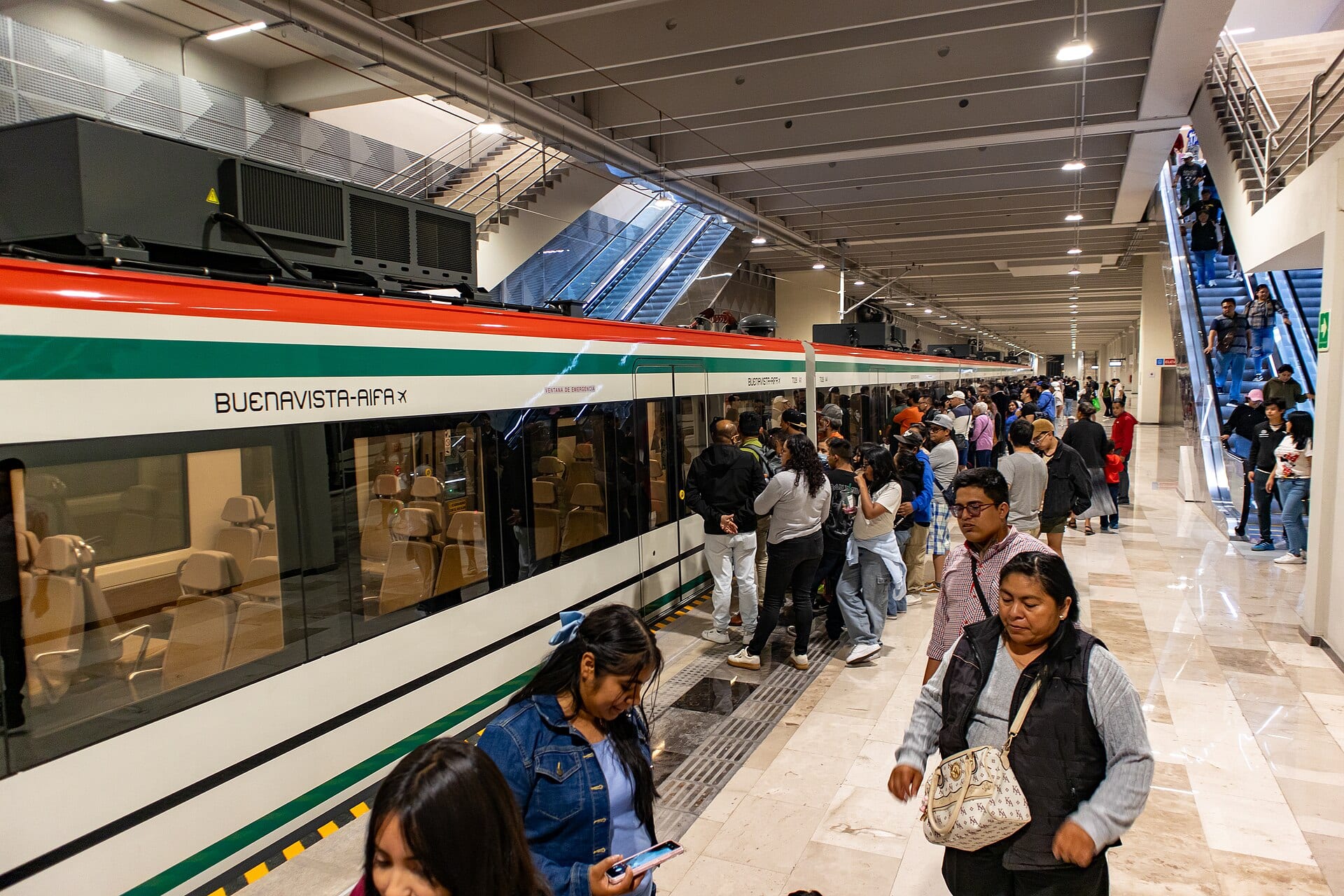 Pasajeros esperan en el andén junto a un tren blanco con franjas roja y verde que dice Buenavista-AIFA.
