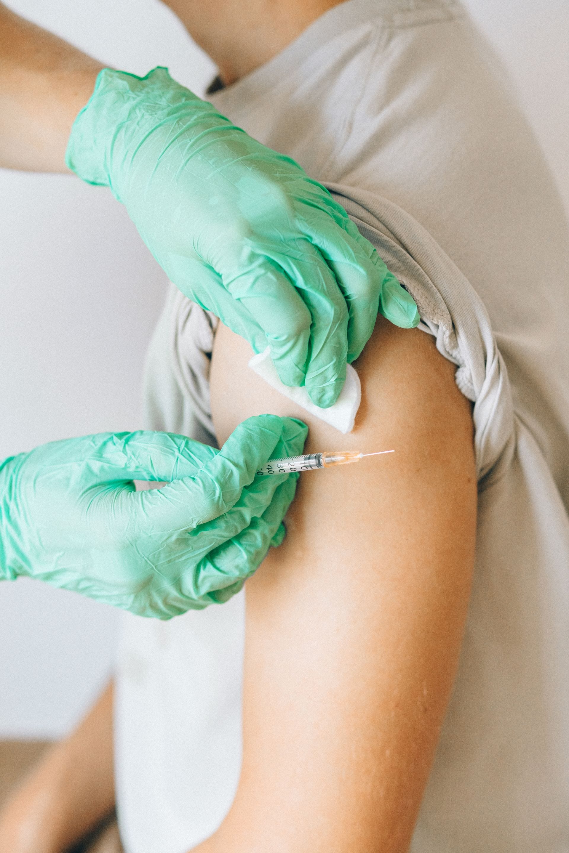 Dos manos con guantes verdes aplican una inyección con una jeringa en el brazo de una persona, mientras sostienen una gasa.