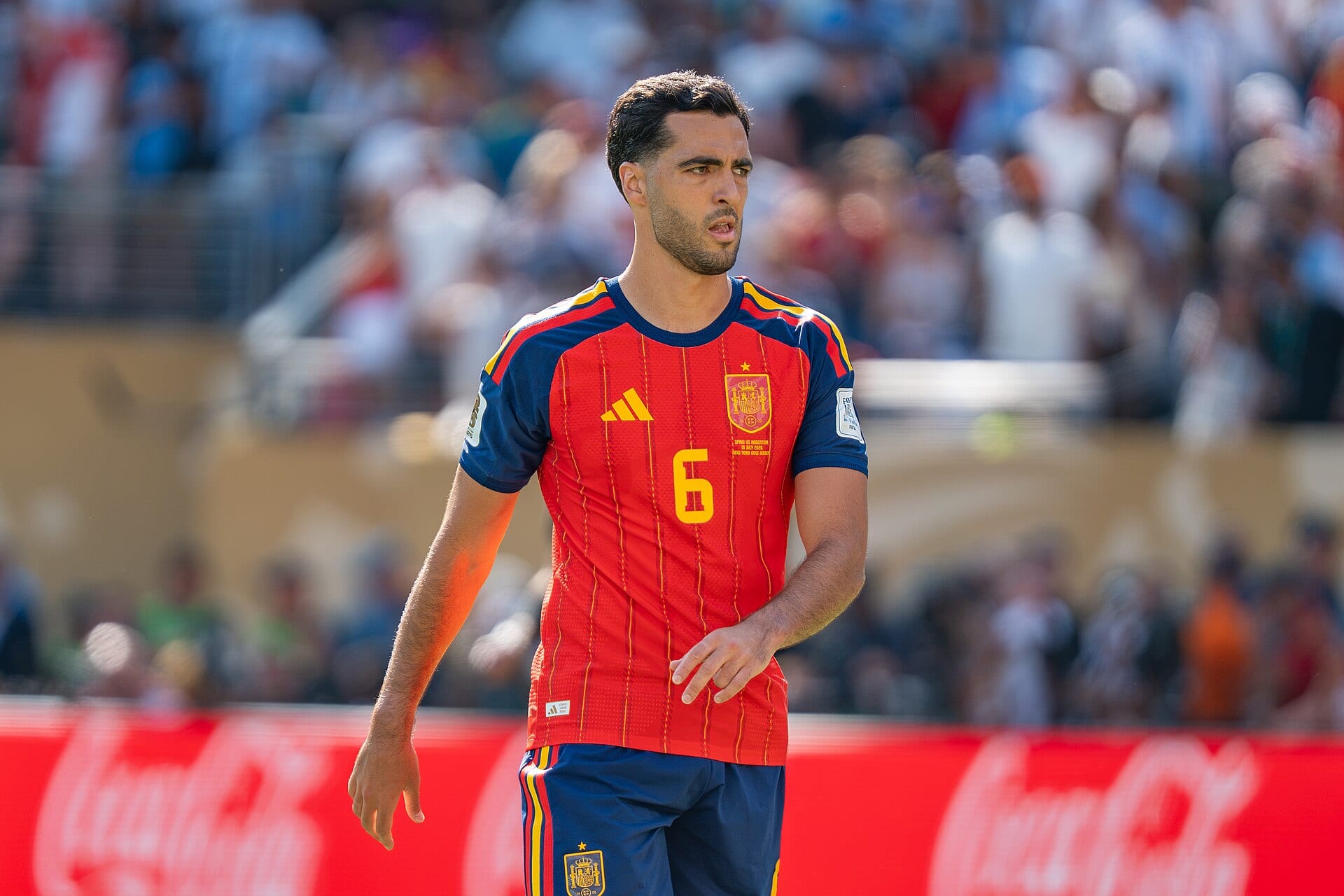 Mikel Merino, con la camiseta roja de España con el número 6, mira hacia un lado durante un partido con la grada borrosa al fondo.