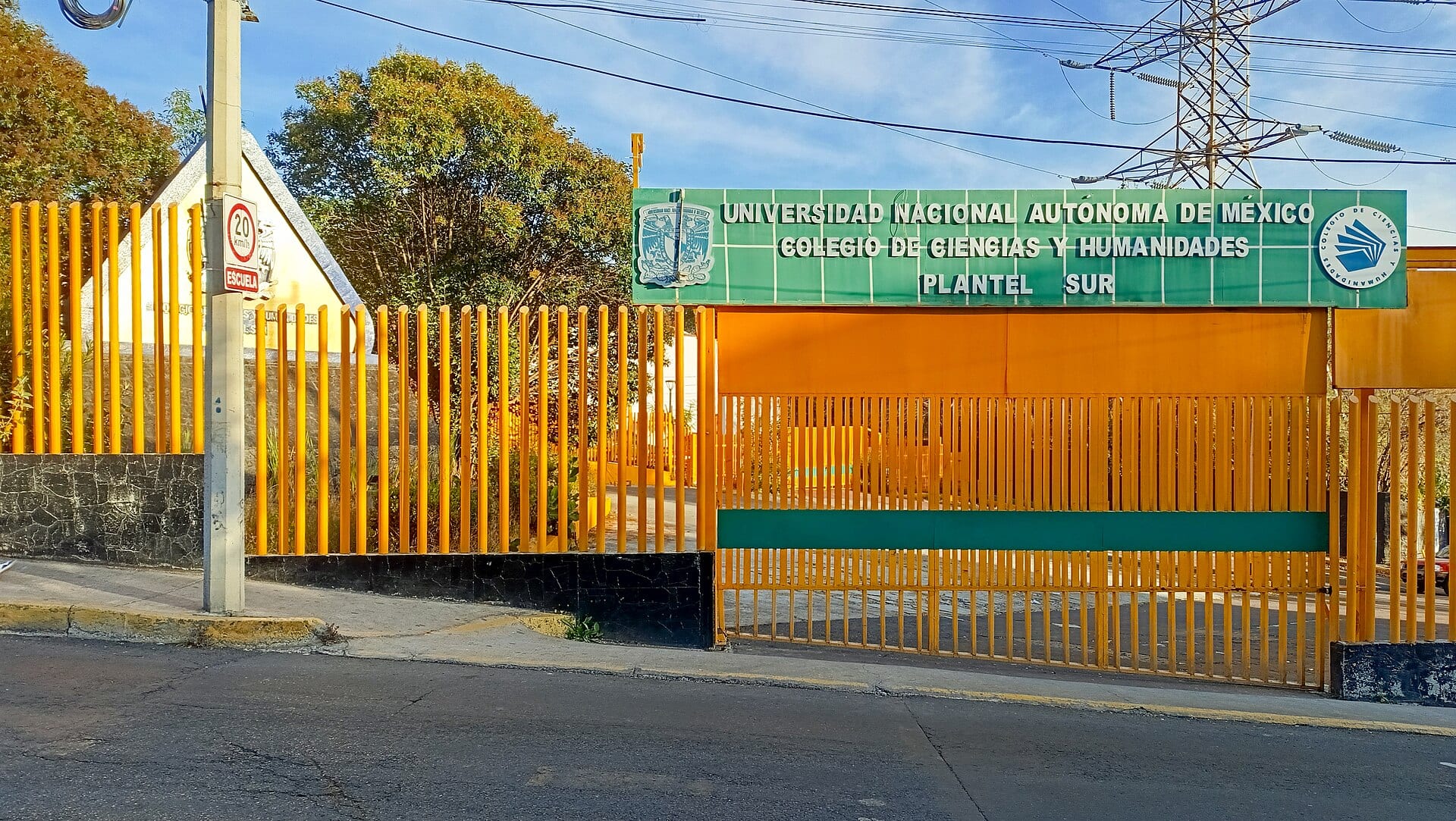 Reja amarilla y letrero verde de la entrada del plantel Sur del Colegio de Ciencias y Humanidades, con un poste y árboles al fondo.