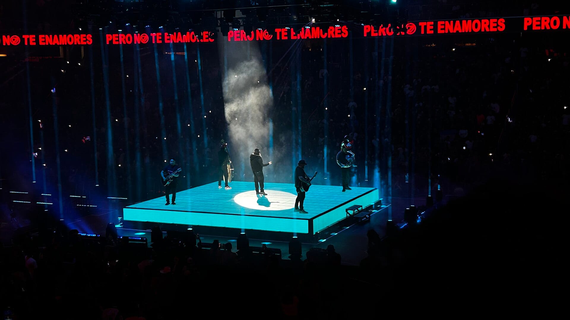 Cinco integrantes de Fuerza Regida tocan sobre una tarima iluminada en turquesa en una arena a oscuras, con un letrero rojo luminoso arriba, en un concierto de 2024.