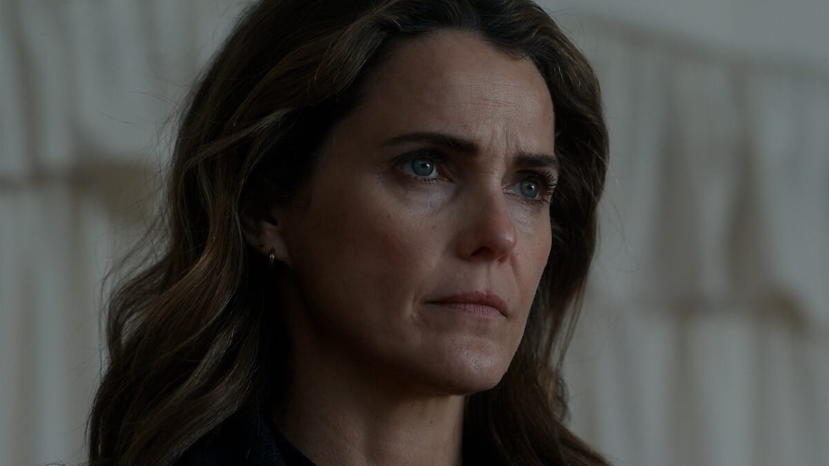 Close-up of a woman with long brown hair and a serious expression, looking to one side in front of a blurred light-colored curtain.