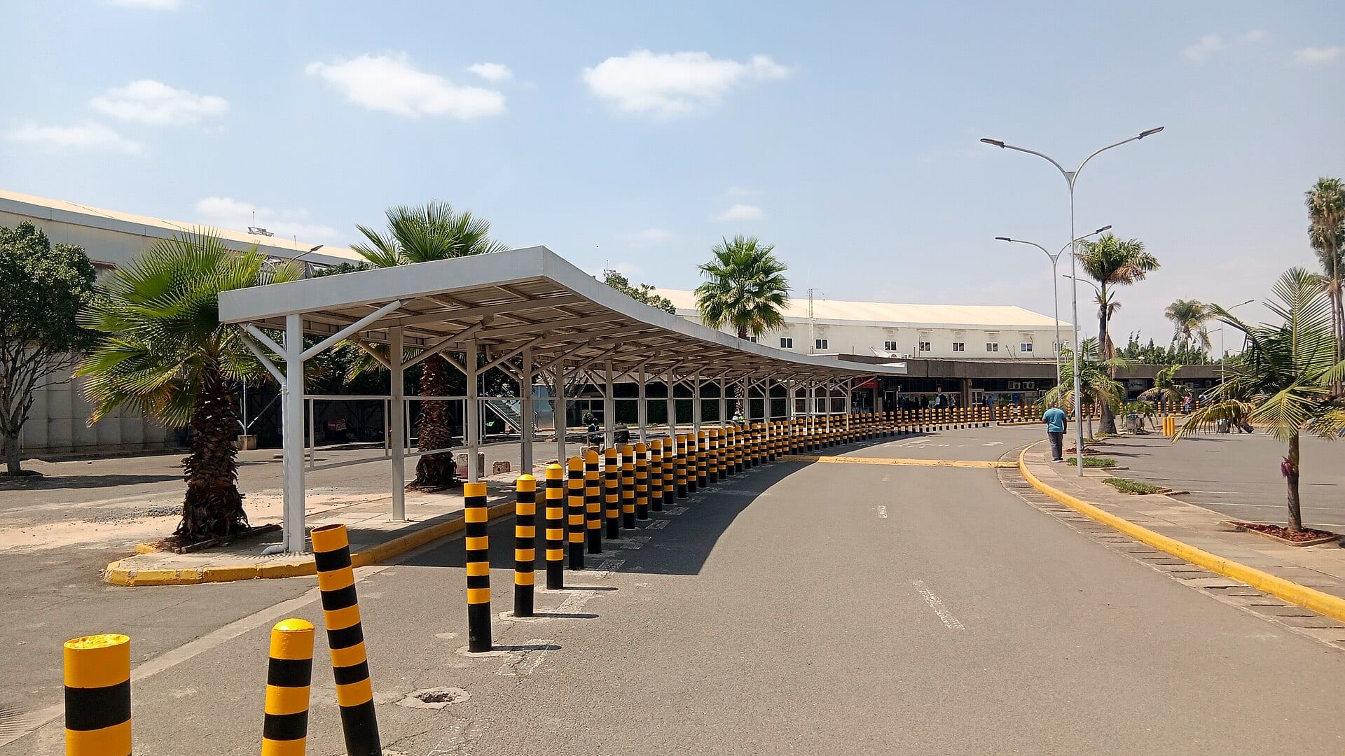Acera cubierta y fila de bolardos amarillos y negros frente al edificio de terminales del aeropuerto de Nairobi.
