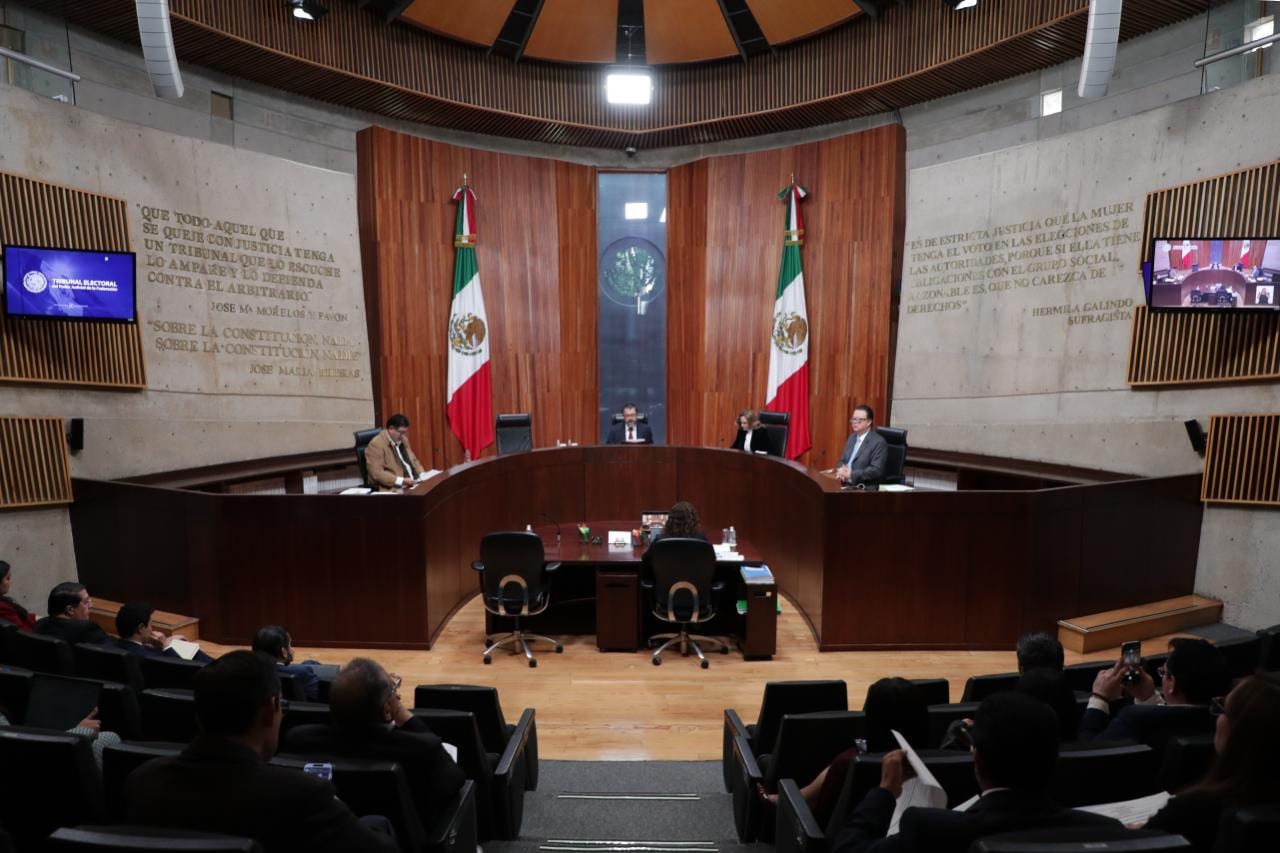 Magistrados en el estrado de la Sala Superior del Tribunal Electoral, entre dos banderas de México, con público sentado en las butacas.