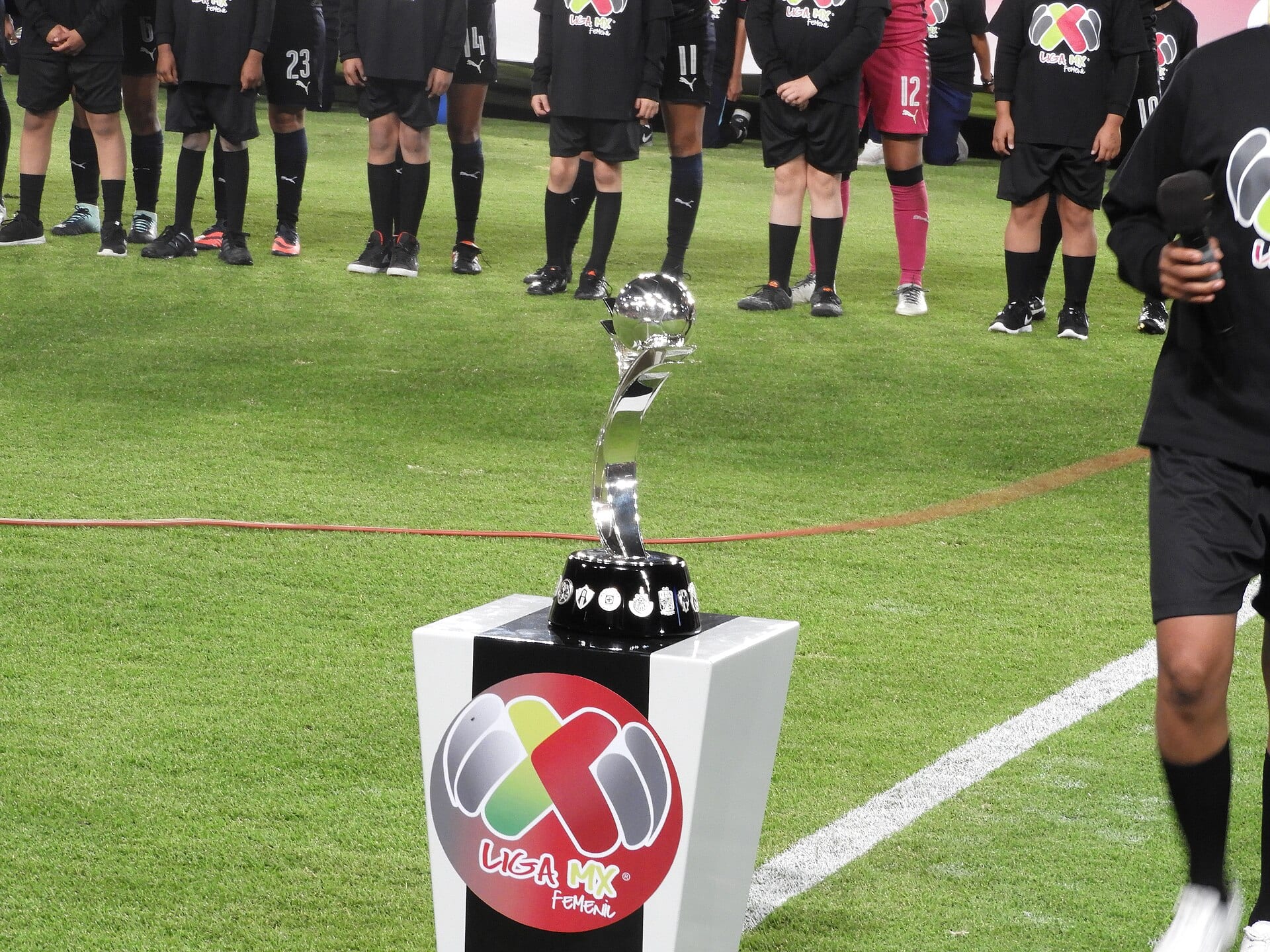El trofeo de la Liga MX Femenil sobre su pedestal, en el césped, con jugadoras y niñas de playera negra al fondo.