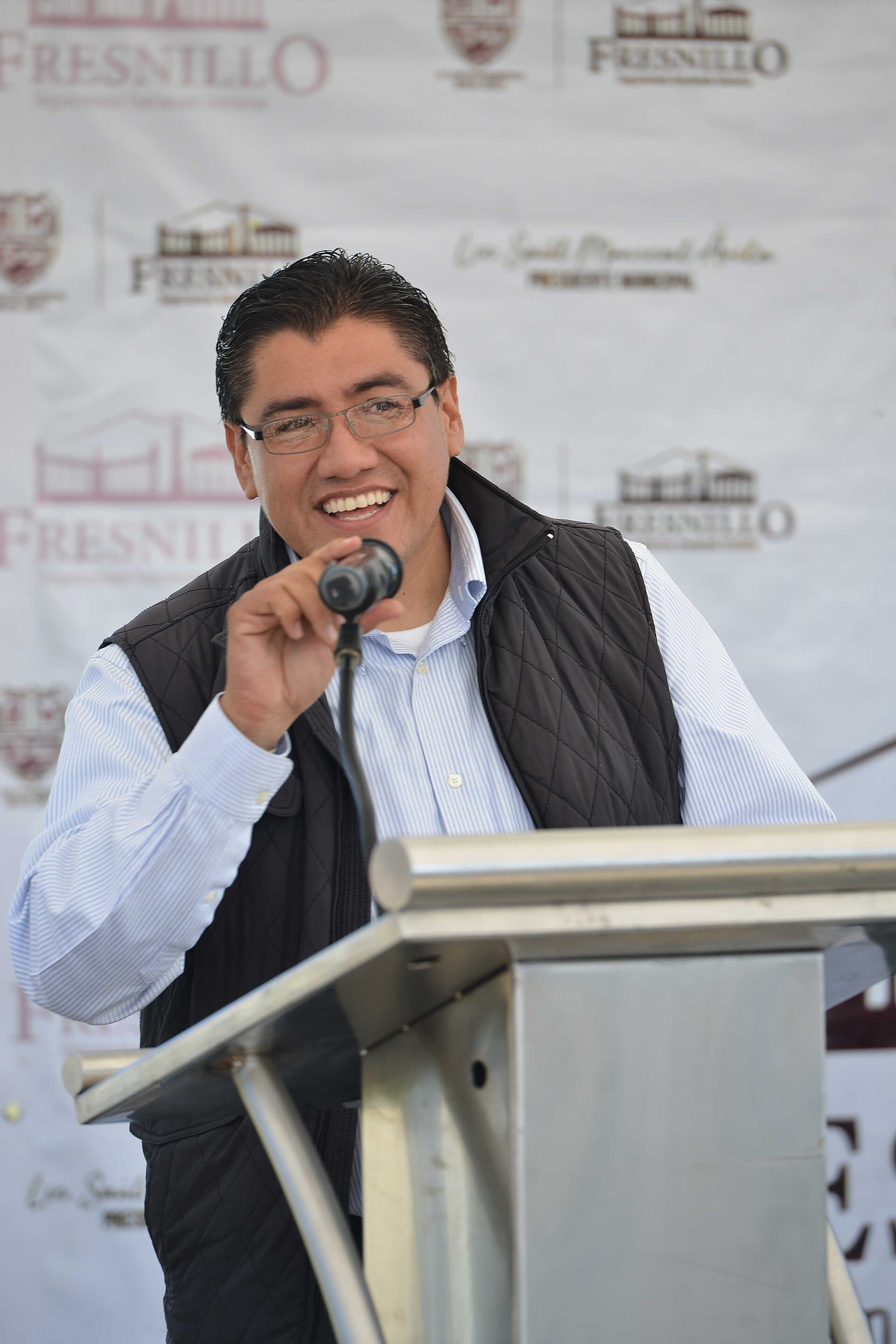 Un hombre de anteojos y chaleco oscuro habla sonriendo ante un micrófono, frente a una lona con el nombre de Fresnillo.
