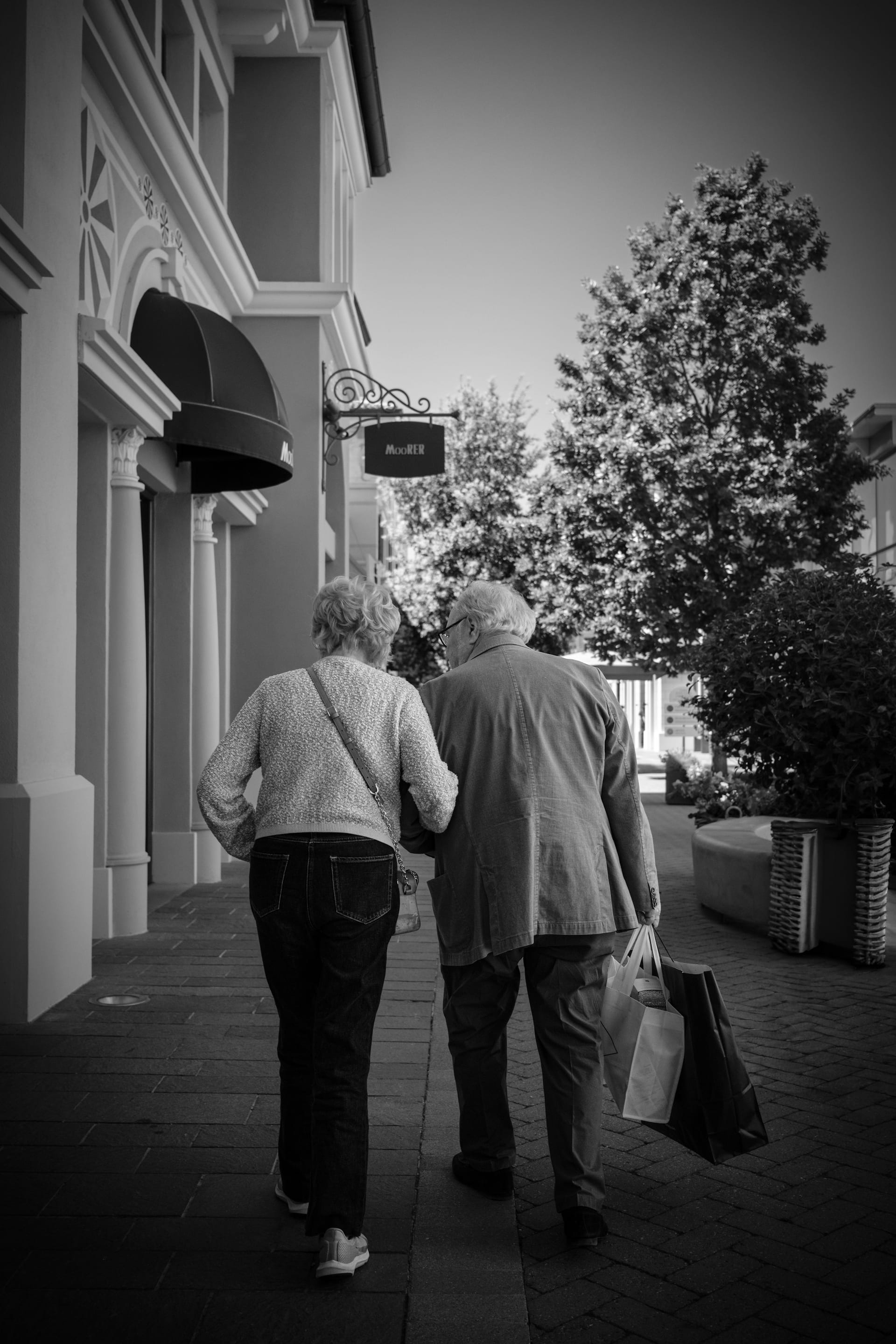 Una pareja de personas mayores camina del brazo, de espaldas, por una banqueta con edificios y árboles, en blanco y negro.