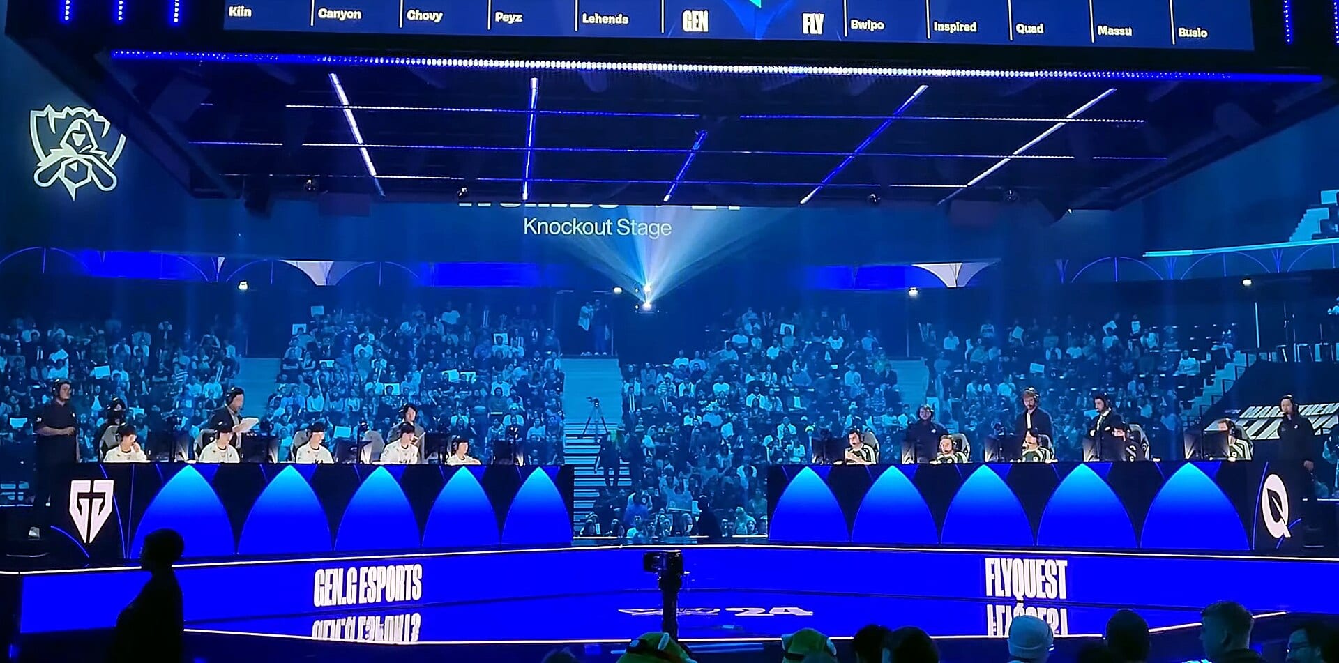 Blue Worlds stage with Gen.G and FlyQuest players seated in front of a full crowd, under a sign that reads Knockout Stage.