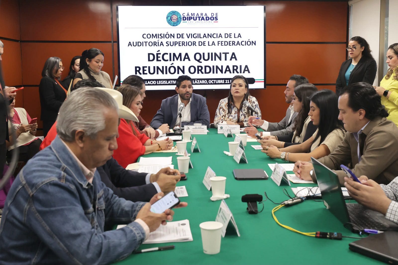 Diputados sentados alrededor de una mesa con mantel verde, bajo una pantalla que dice "Décima Quinta Reunión Ordinaria" de la Comisión de Vigilancia.