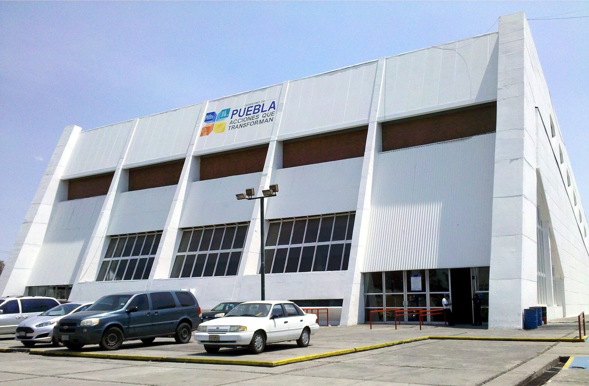 Fachada blanca e inclinada de un gimnasio con el logotipo del Gobierno de Puebla, y varios autos estacionados al frente.