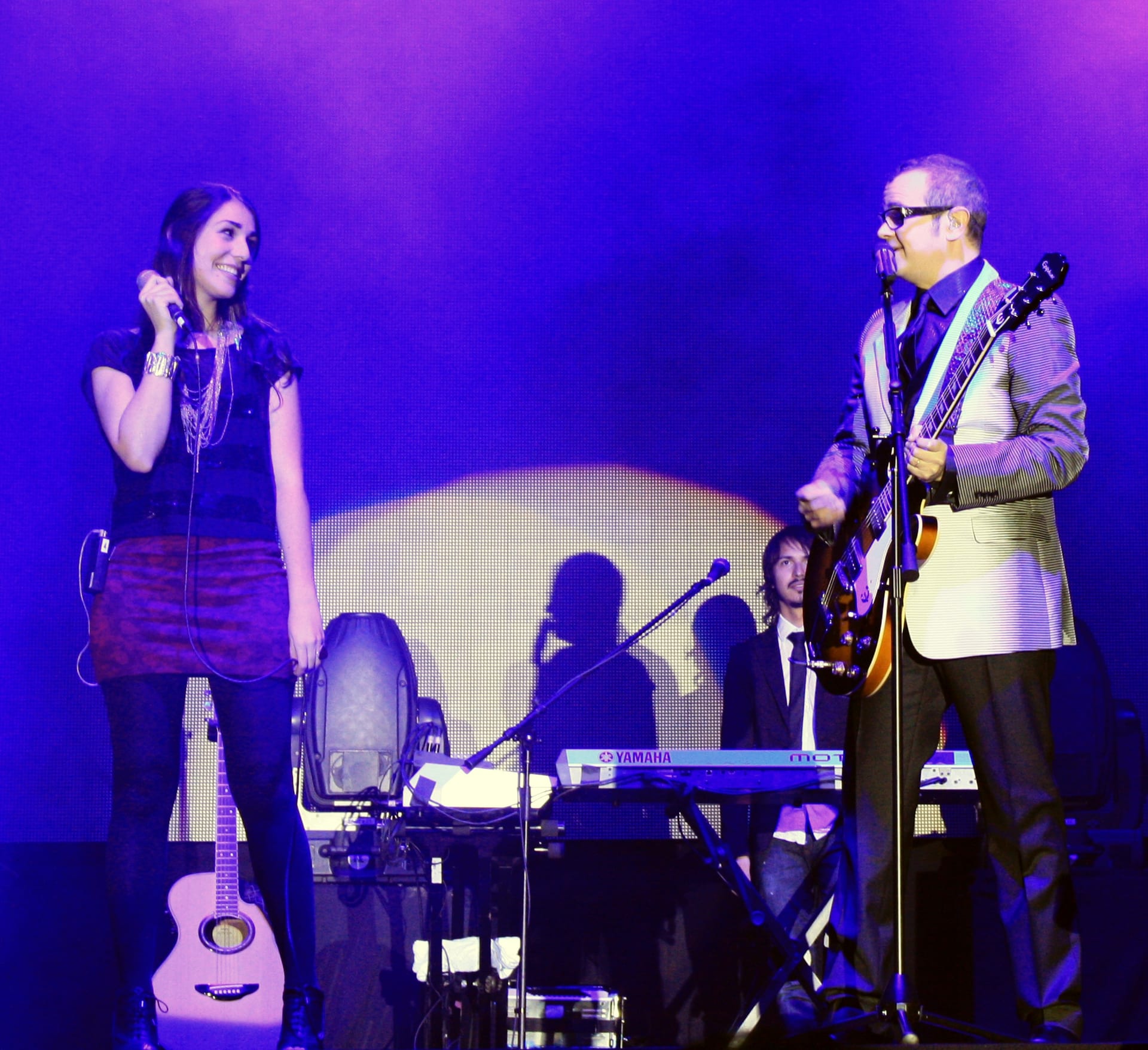 El cantante Aleks Syntek, con lentes y saco a rayas, toca la guitarra y canta junto a una corista, en un concierto con luces moradas.