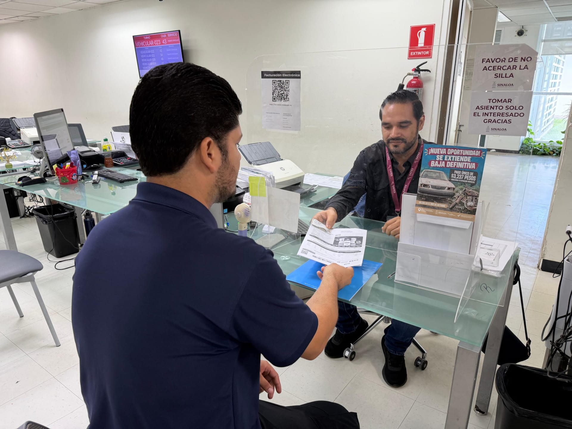 Un contribuyente de playera azul marino entrega un recibo a un empleado del SATES con gafete, detrás de una mampara de acrílico en una ventanilla de atención.