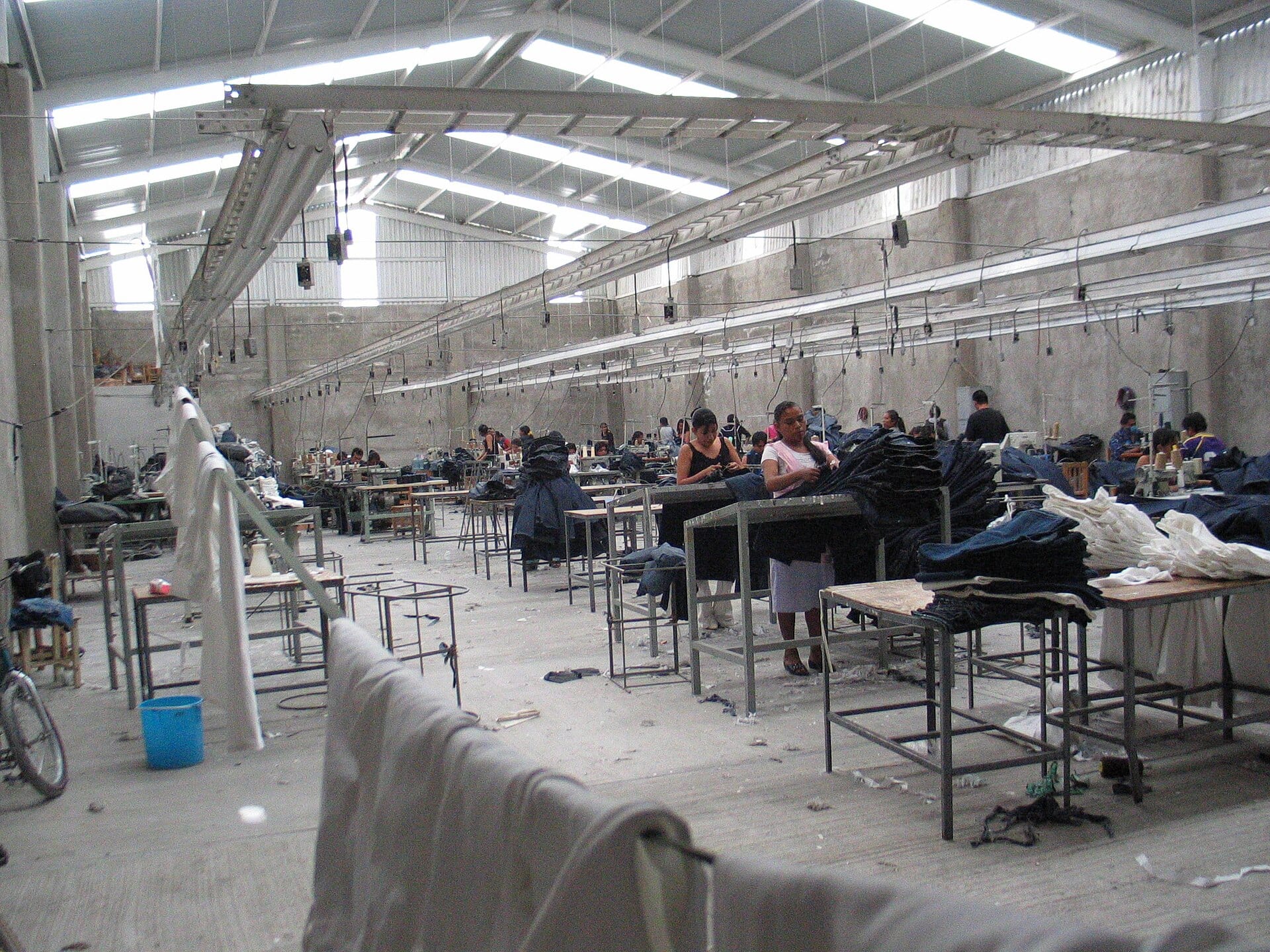 Women stand at tables stacked with denim garments inside a large factory hall with a corrugated roof and skylights.