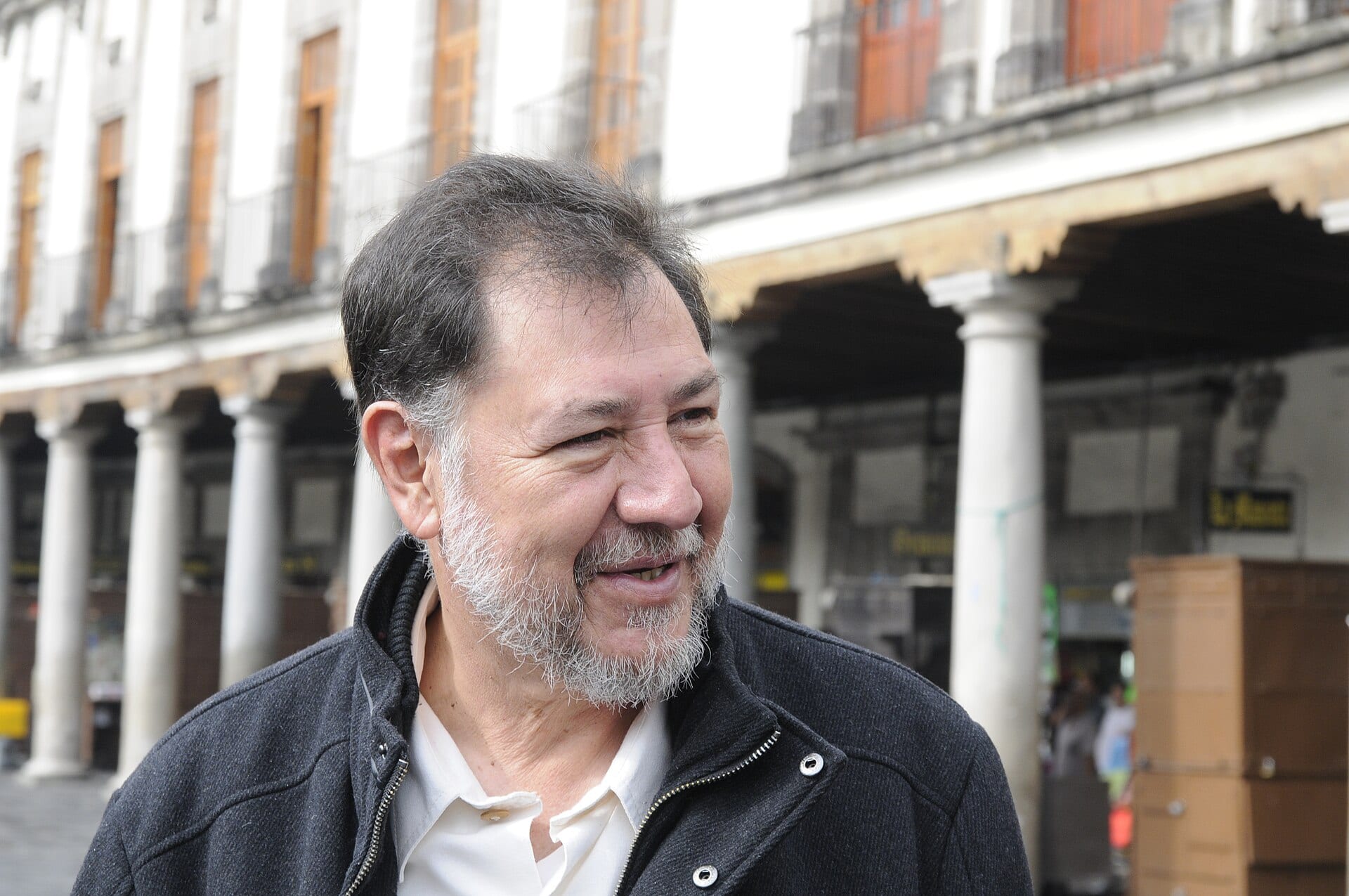 Gerardo Fernández Noroña, de barba cana y chamarra oscura, sonríe mirando hacia un lado frente a unos portales de columnas blancas.