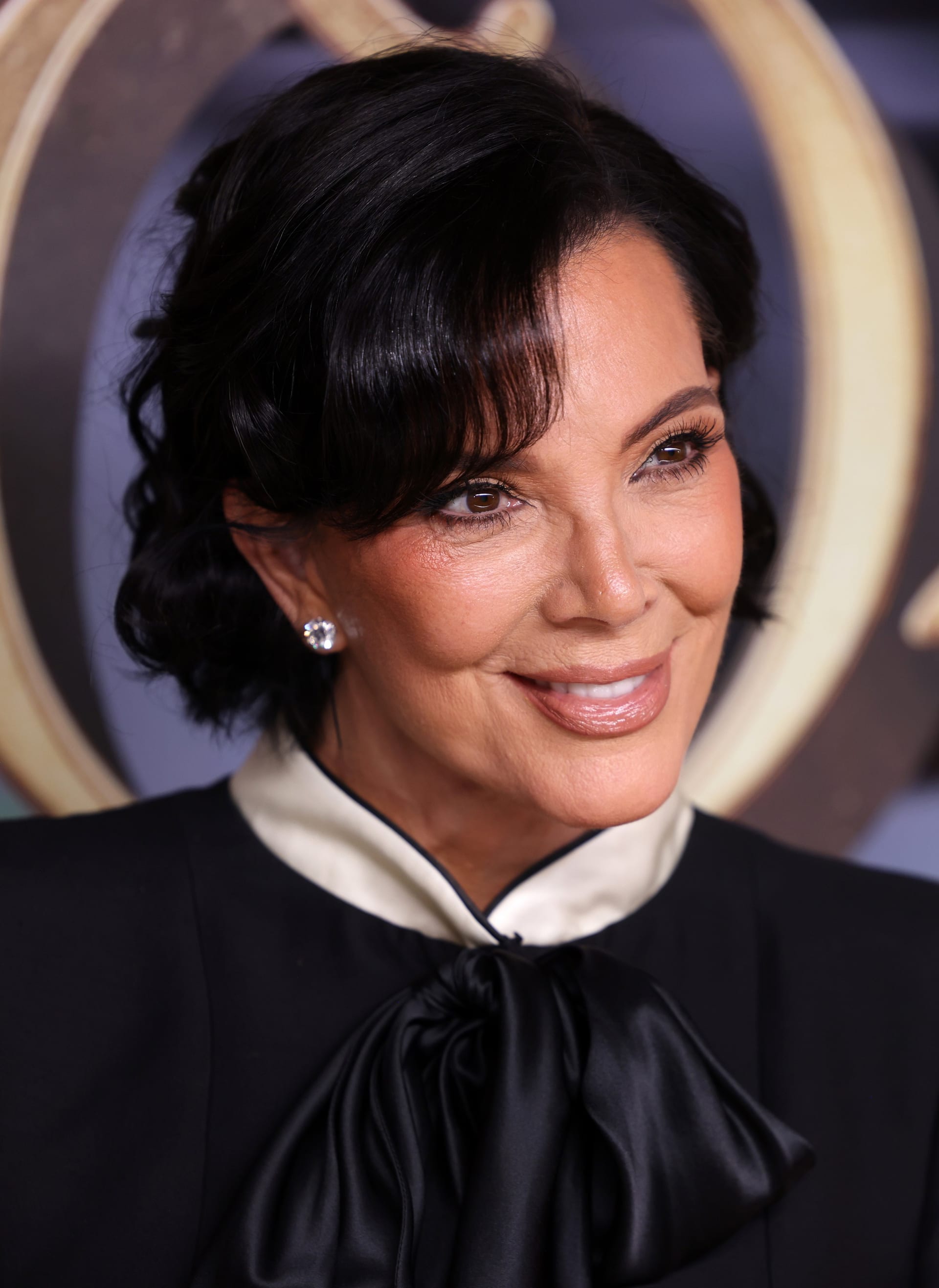 Kris Jenner, de pelo oscuro recogido y blusa negra con moño de satén, sonríe de lado frente a un fondo dorado.