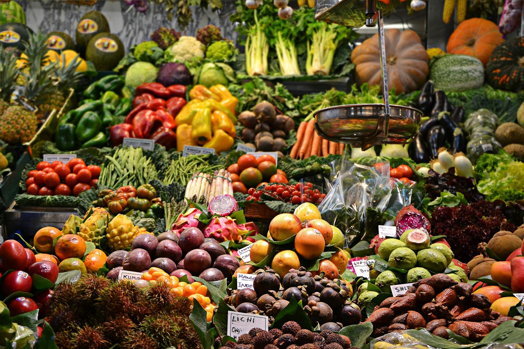 Puesto de mercado con montones de frutas y verduras de colores, entre ellas pimientos, tomates, calabazas, pitahayas y litchis, con etiquetas de precio.