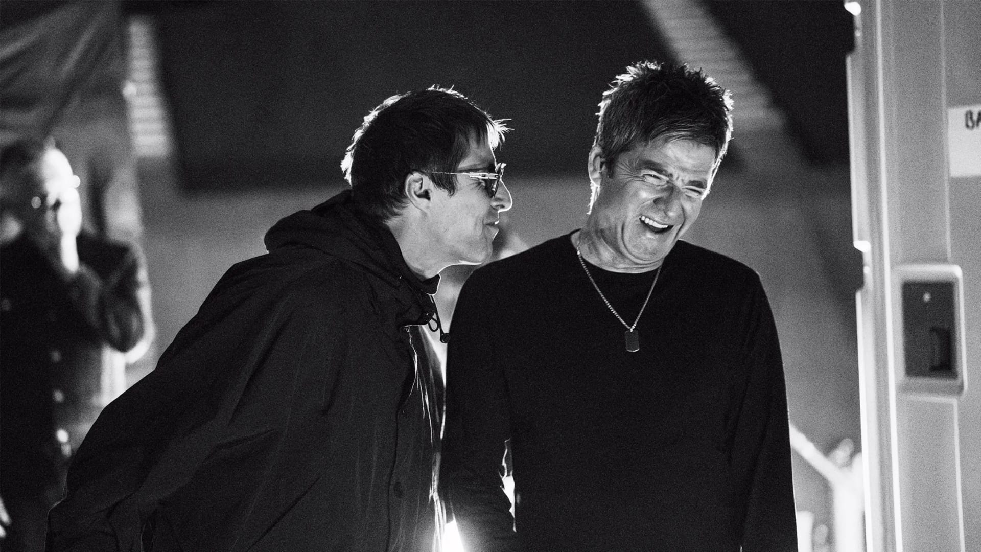 Liam y Noel Gallagher, en blanco y negro, conversan entre bastidores; Liam, con lentes oscuros, se inclina hacia Noel, que se ríe a carcajadas.