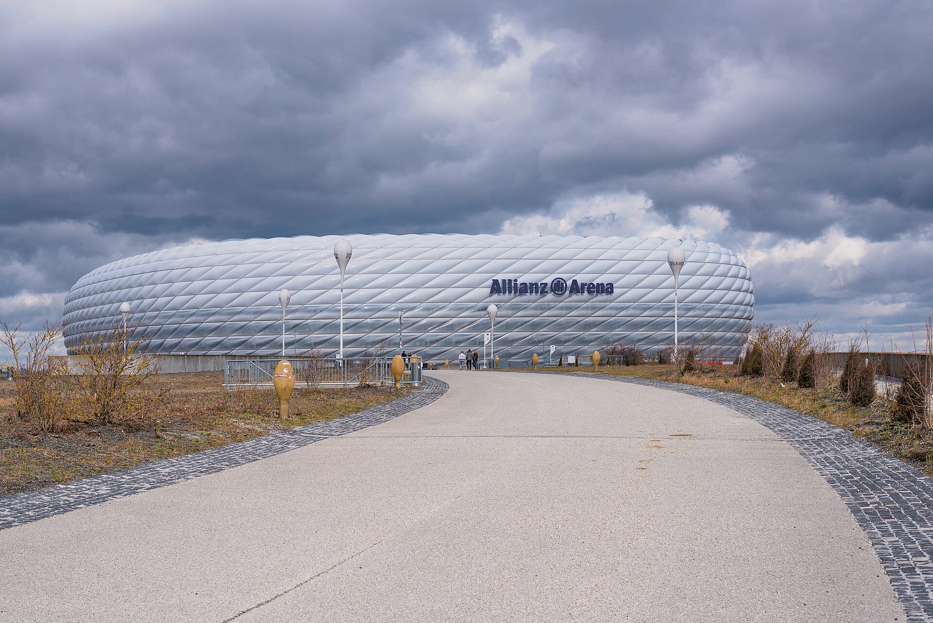 Fachada blanca del Allianz Arena con su rótulo, vista desde un camino de concreto bajo un cielo de nubes oscuras.