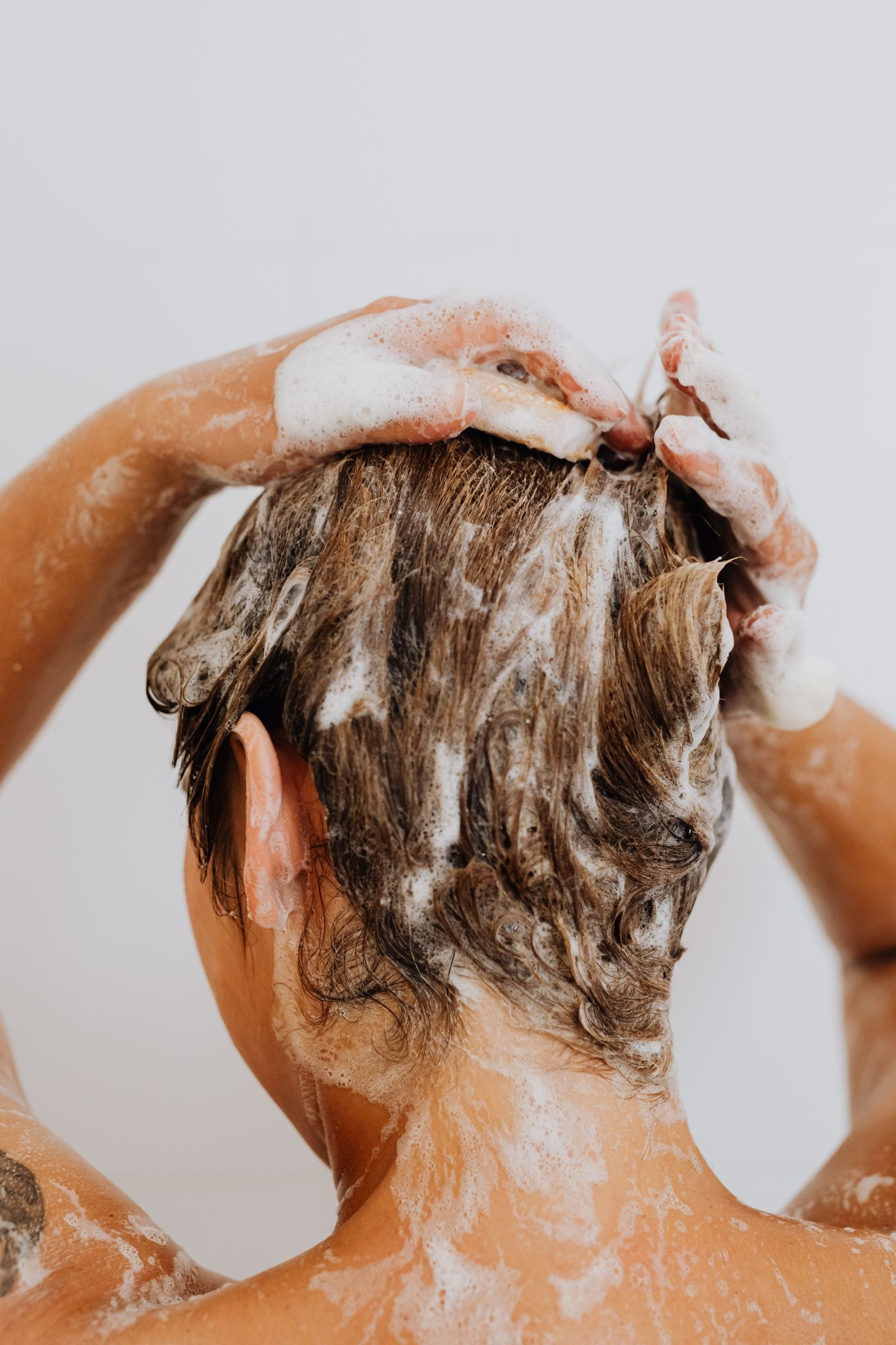 Una persona vista de espaldas se enjabona el cabello con espuma de champú bajo la ducha.