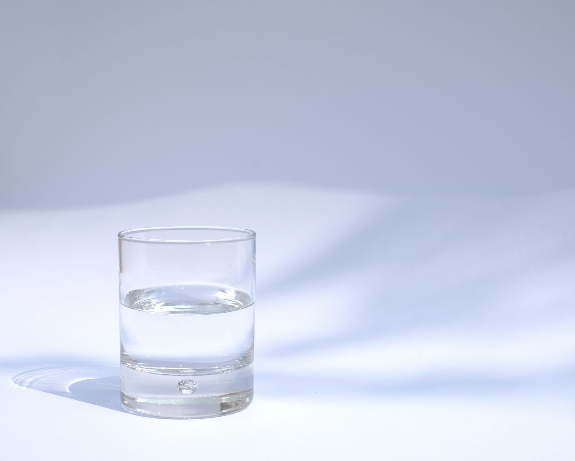 Un vaso transparente con agua hasta la mitad sobre una superficie blanca, con sombras azuladas de fondo.