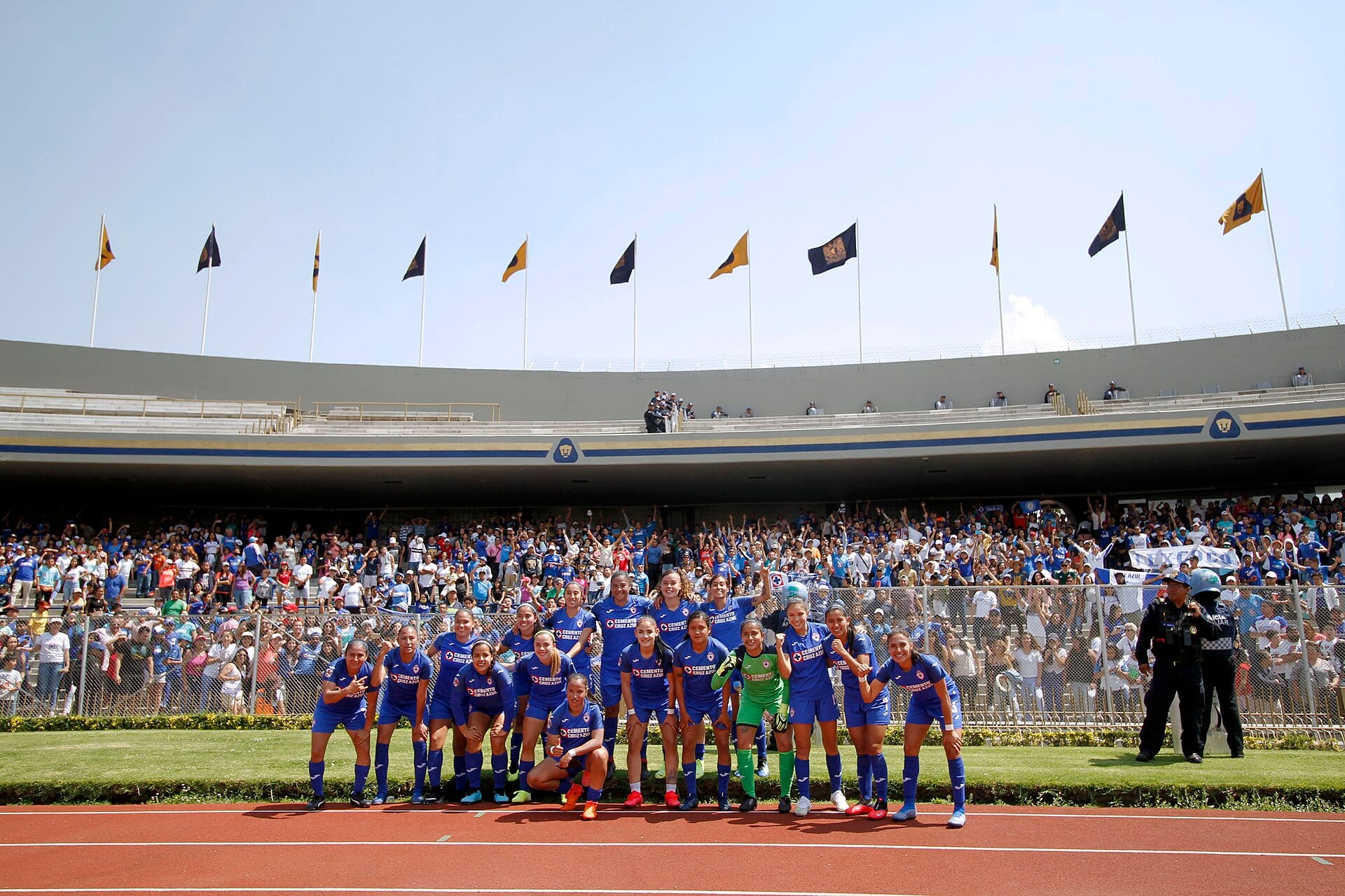Jugadoras de Cruz Azul con uniforme azul posan de pie ante las gradas llenas de aficionados, con banderas universitarias ondeando al fondo.