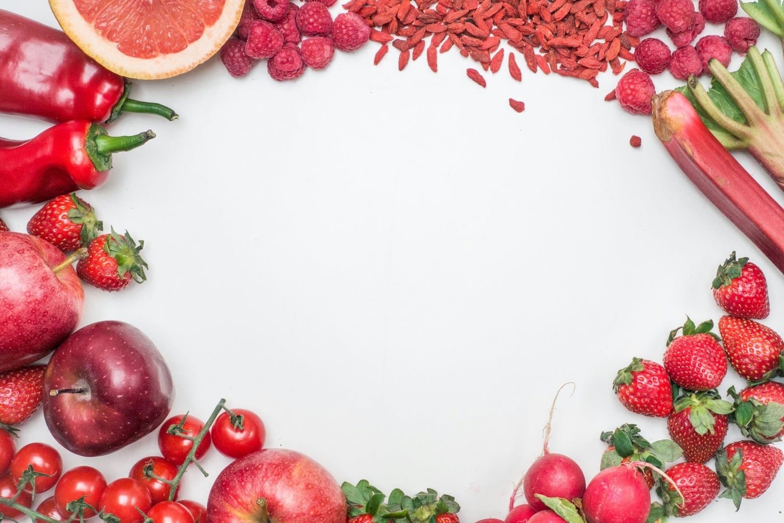 Vista cenital de manzanas, fresas, jitomates, rábanos y chiles rojos repartidos en el borde de una superficie blanca.