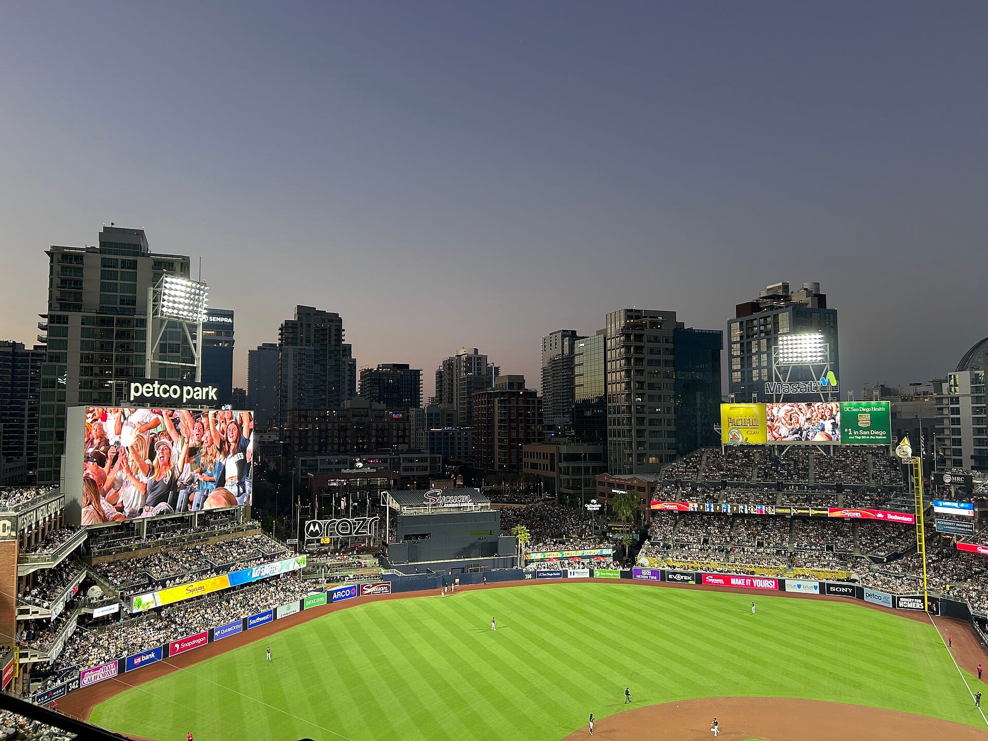 Vista del campo verde de Petco Park al anochecer, con las gradas llenas, las pantallas gigantes y los edificios del centro de San Diego al fondo.