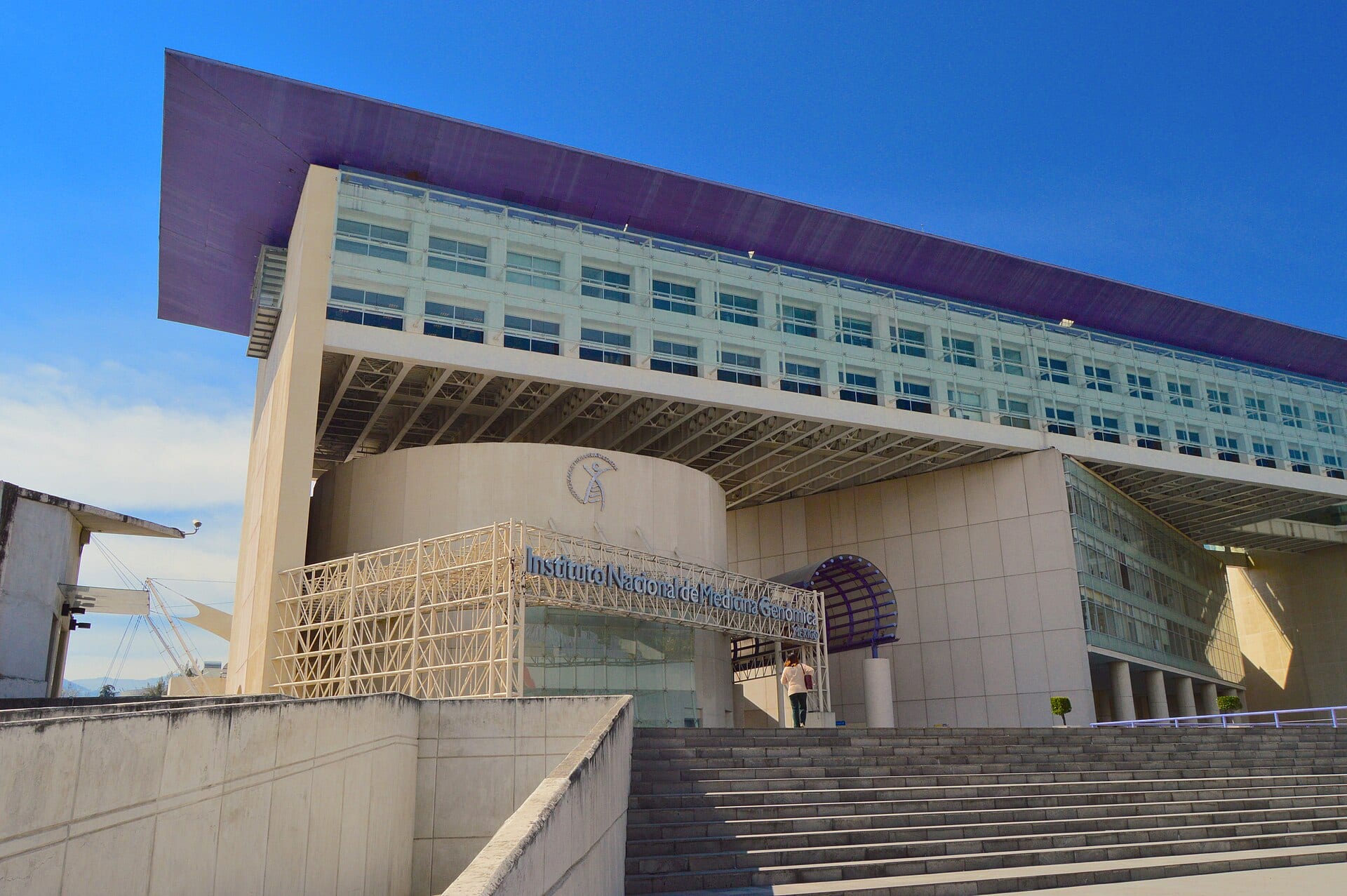 Fachada del Instituto Nacional de Medicina Genómica, un edificio de cristal y concreto con techo morado y una escalinata al frente.