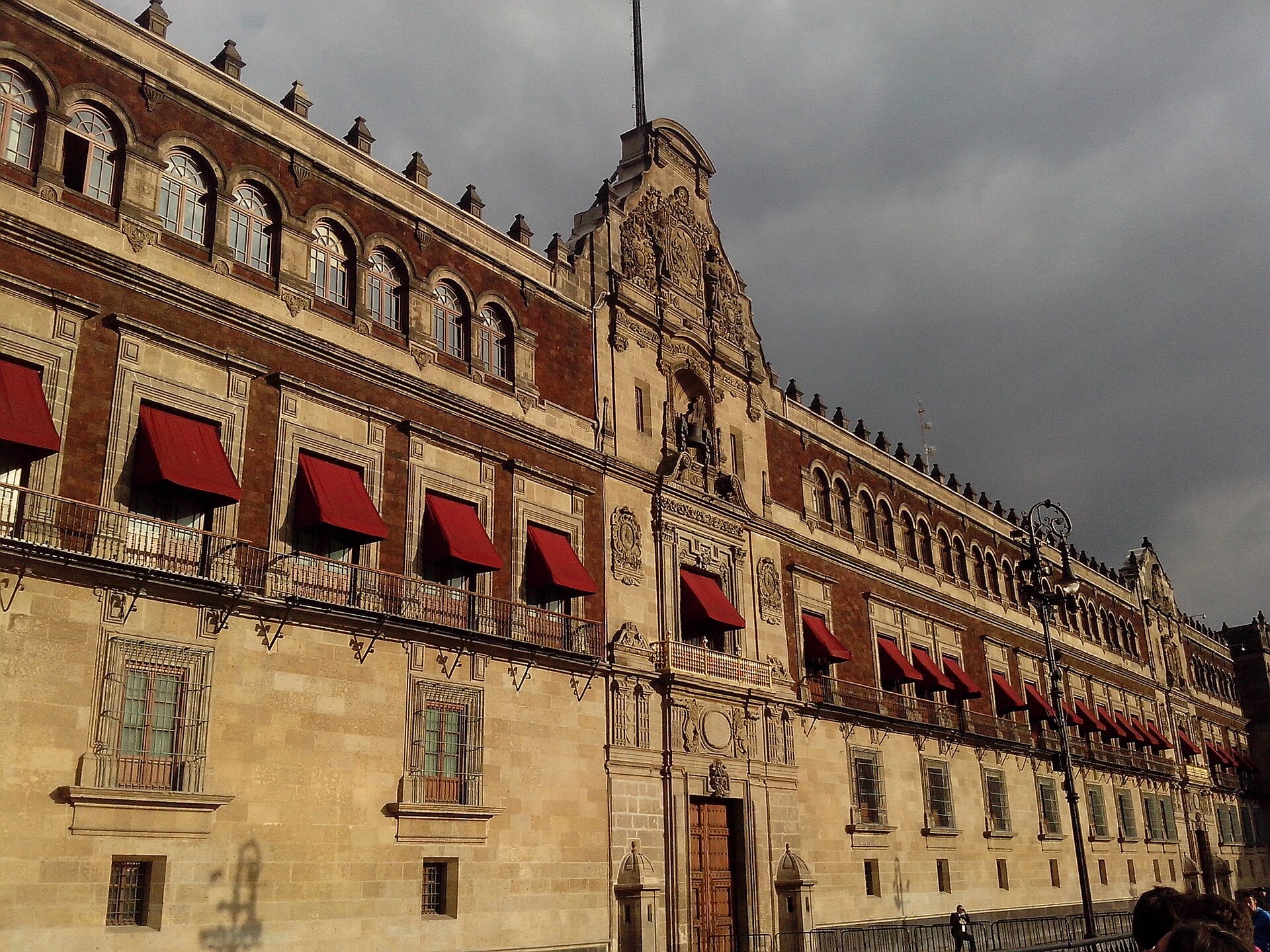 Fachada de piedra de tres niveles de un edificio colonial, con balcones cubiertos por toldos rojos y un remate ornamentado al centro, bajo un cielo nublado.