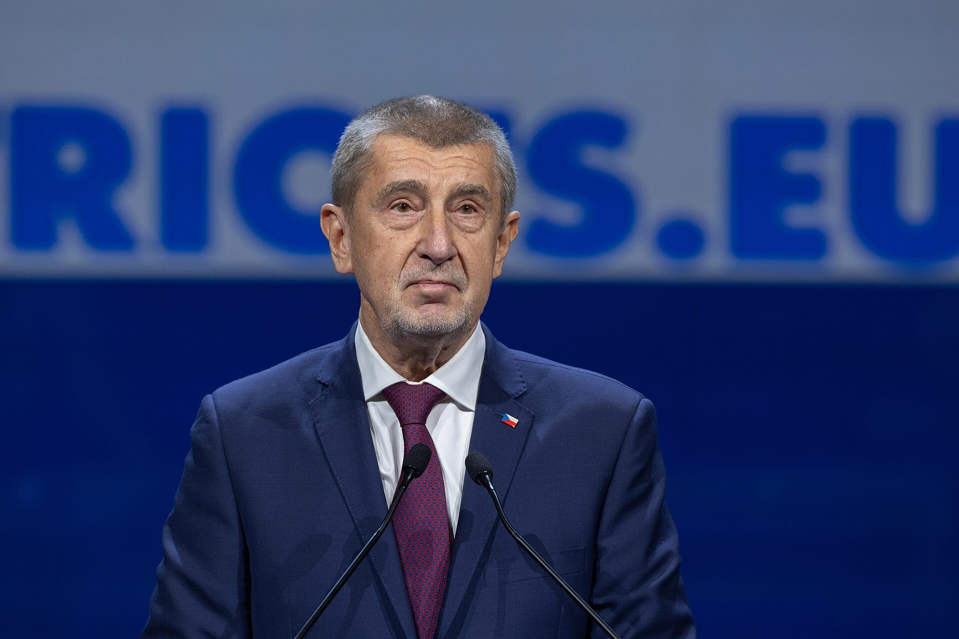 Andrej Babiš, de traje azul marino y corbata granate, habla frente a dos micrófonos ante un fondo azul con letras grandes.