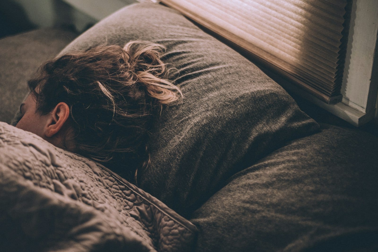 Una mujer duerme de lado sobre una almohada gris, con luz natural entrando por una persiana.