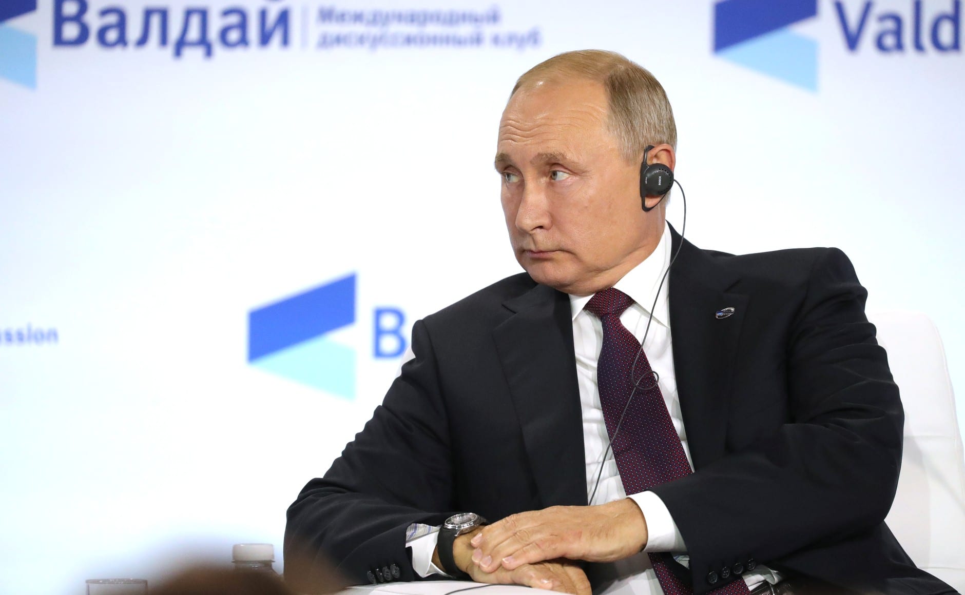 Vladimir Putin, de traje oscuro y con un auricular, mira hacia un lado con las manos cruzadas frente a un fondo blanco con el logotipo del Club Valdai.