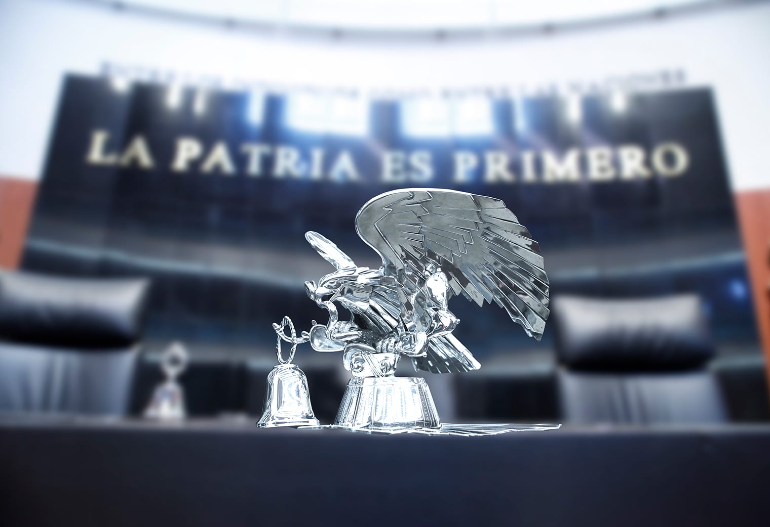 Águila de plata y campana sobre la mesa de la presidencia del Senado, con el lema "La patria es primero" desenfocado al fondo.