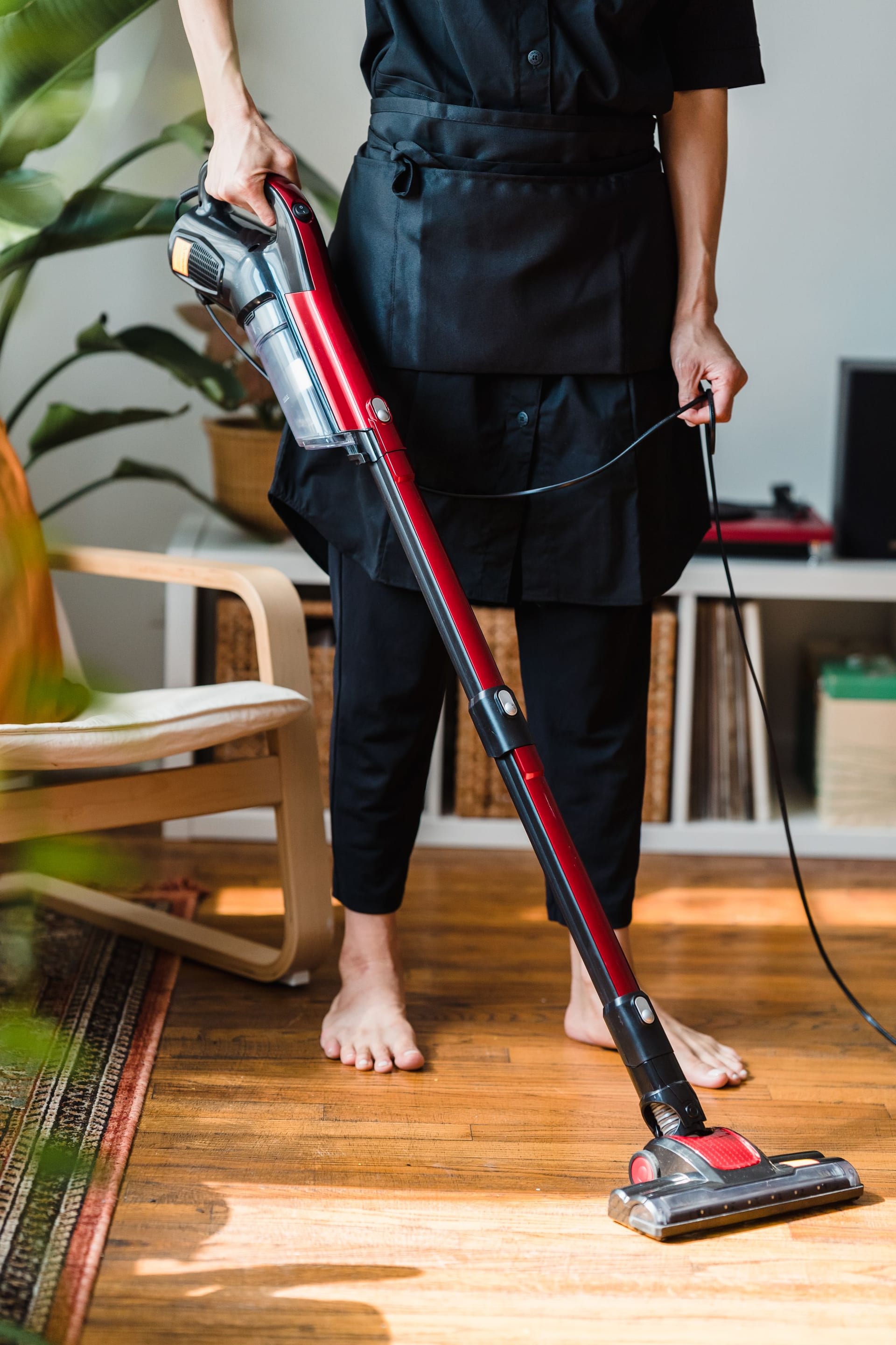 Una persona descalza pasa una aspiradora de mano roja y gris sobre un piso de madera, en una sala con plantas.