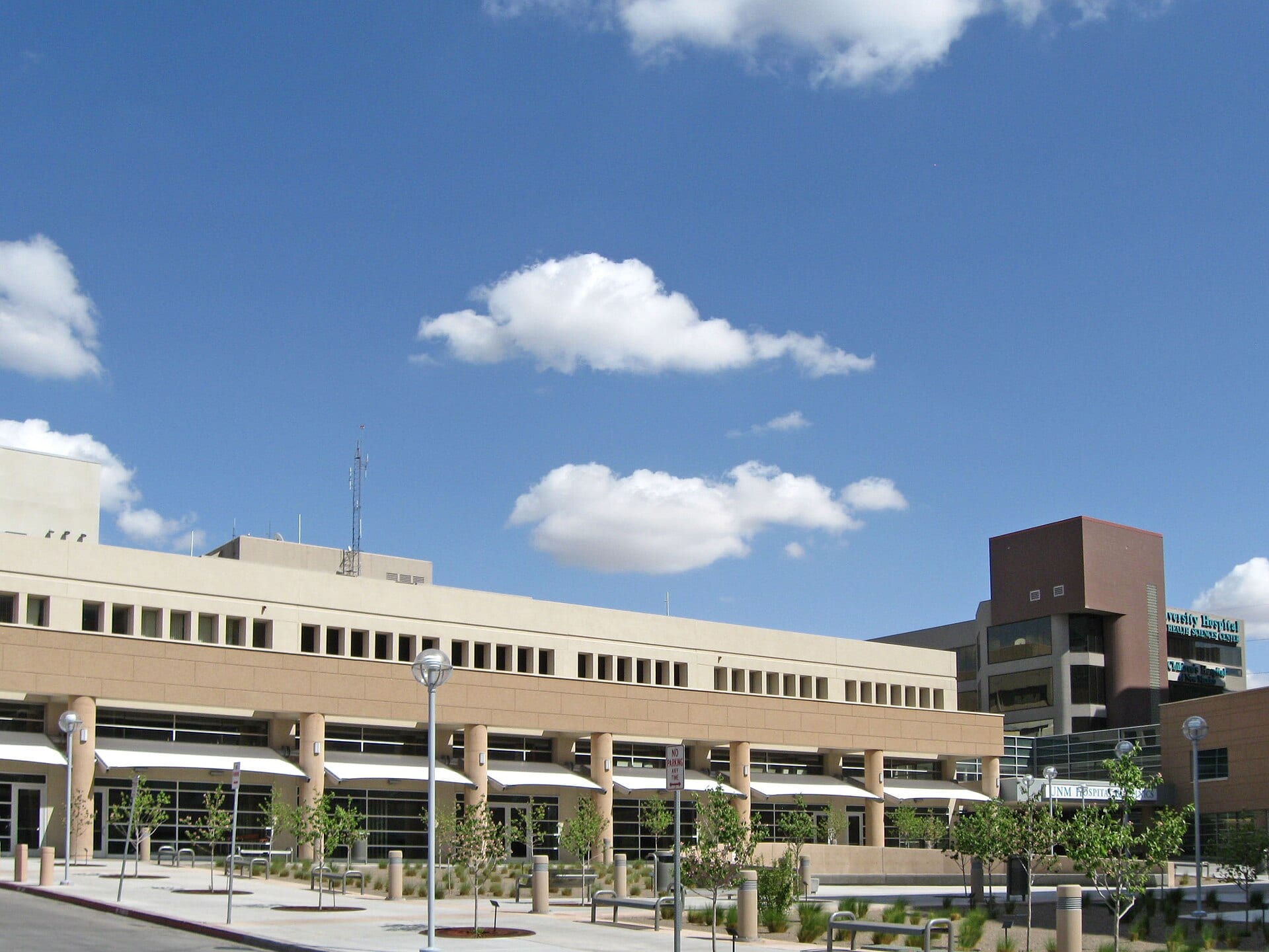 A beige, multistory hospital building with columns, white awnings and young trees in the plaza, under a blue sky with clouds.