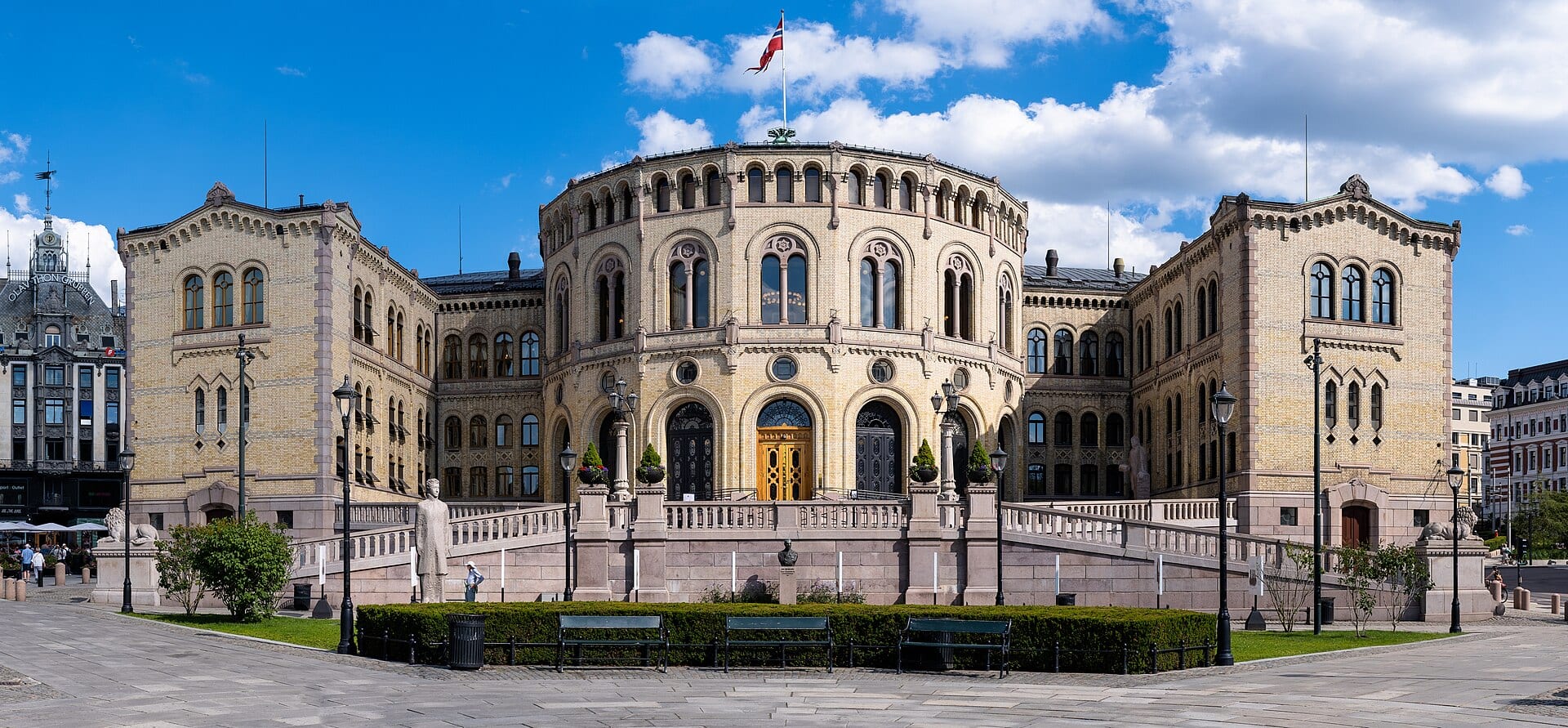 Fachada de ladrillo claro del Parlamento noruego en Oslo, con un cuerpo central curvo, puertas arqueadas, una bandera en lo alto y una plaza al frente.